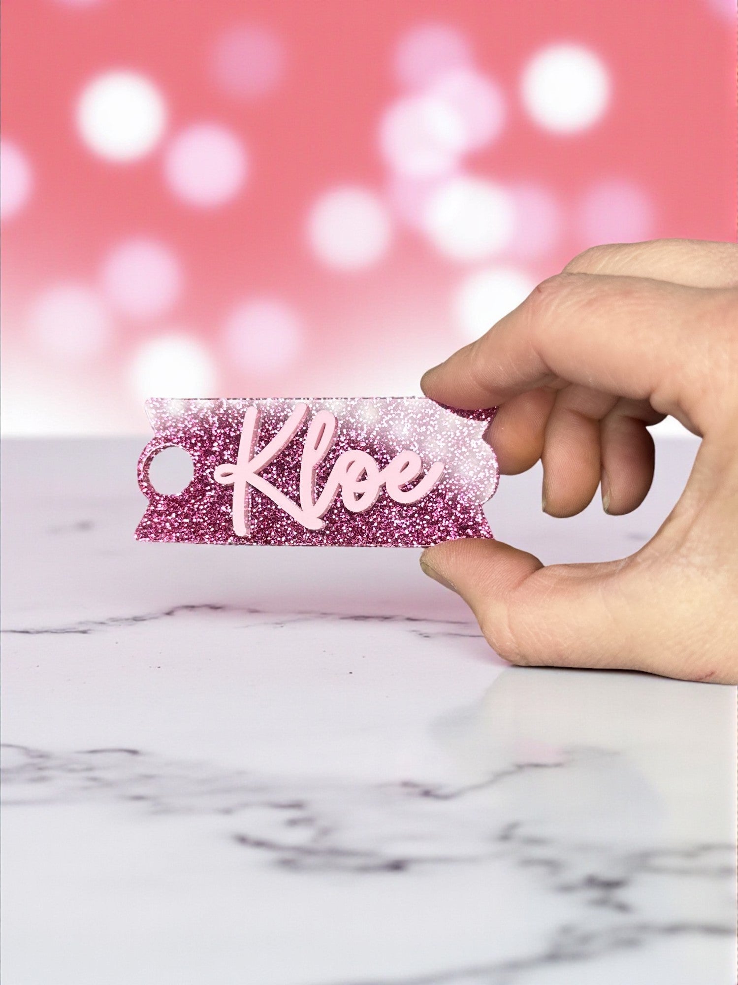 A hand is holding a pink glitter keychain with the name "Kloie" written on it.