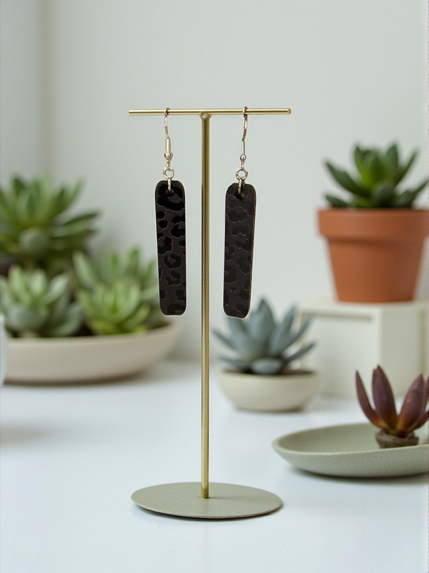 A pair of black leopard print earrings is displayed on a gold stand, with potted plants and other decorative items in the background.