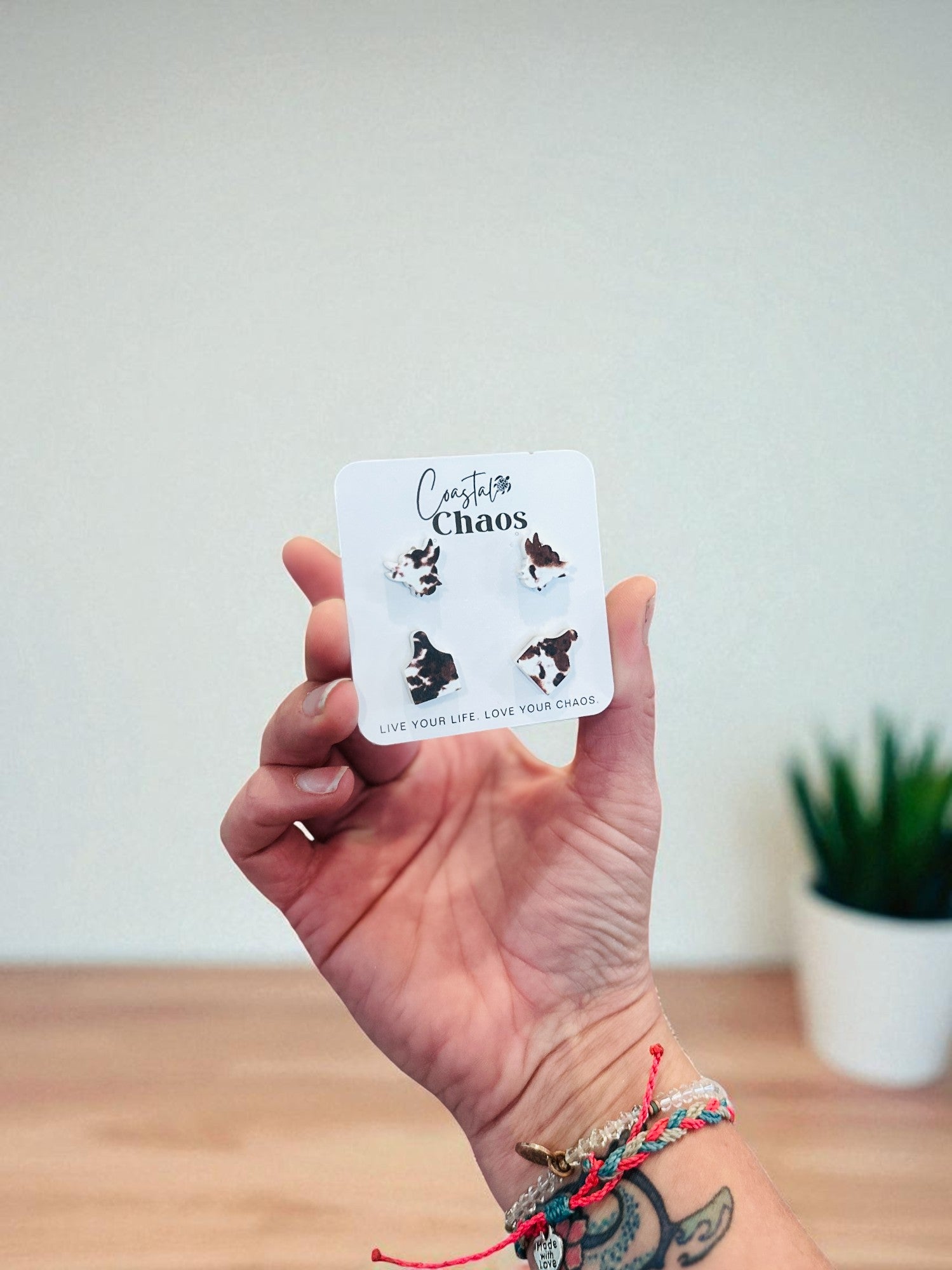 A hand holding a small white card with earrings and text on it, against a plain white wall.