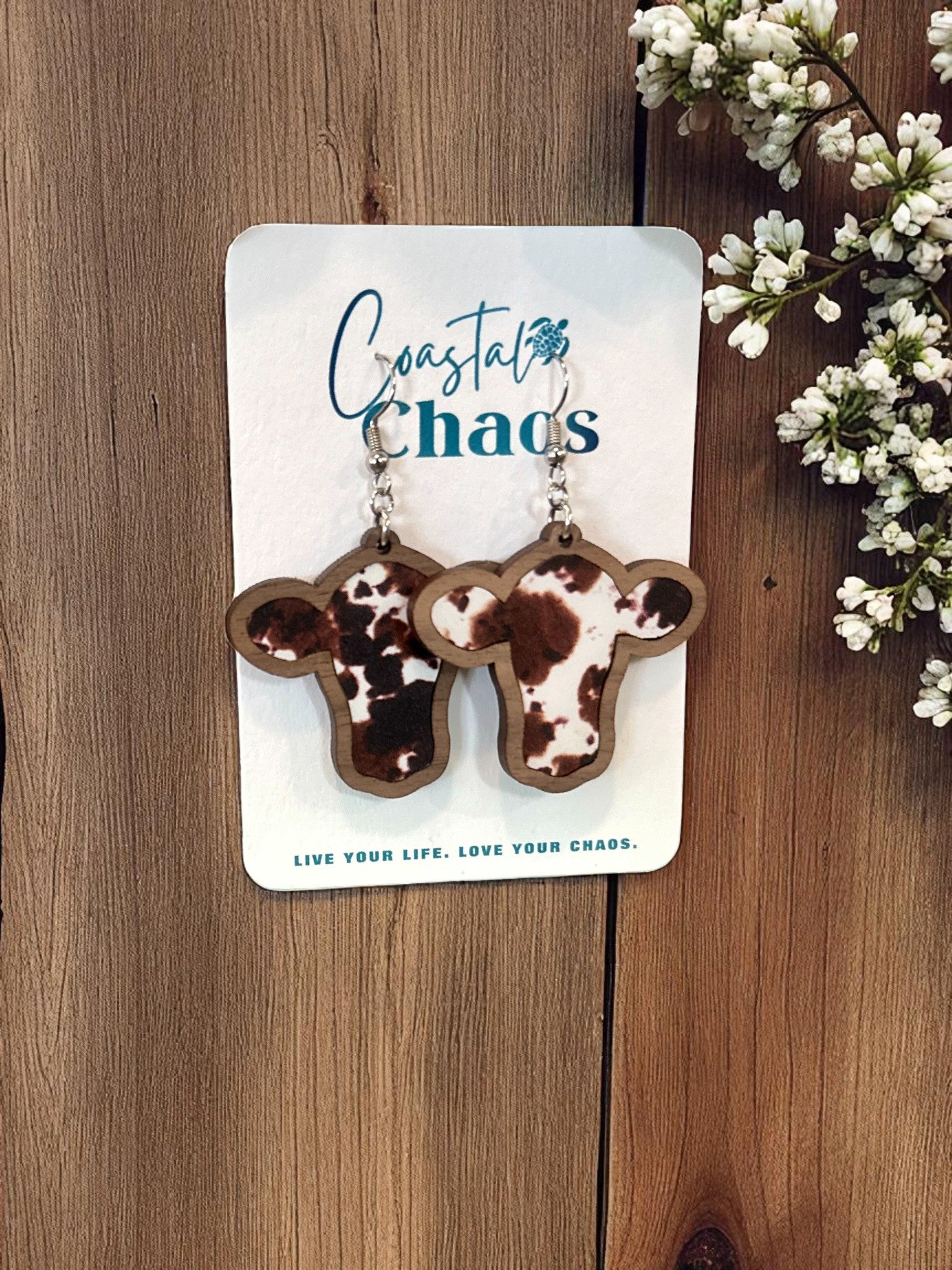 A pair of wooden earrings shaped like cow heads, hanging on a wooden surface with a floral arrangement to the right.