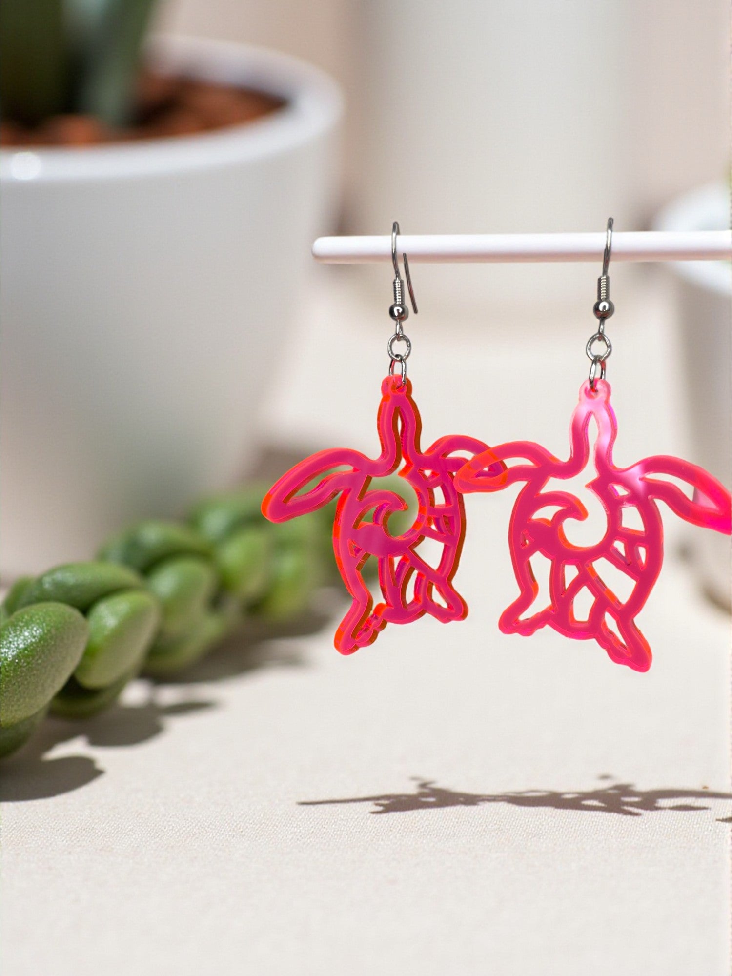 A pair of pink turtle-shaped earrings hanging from a white earring hook, with green plants in the background.