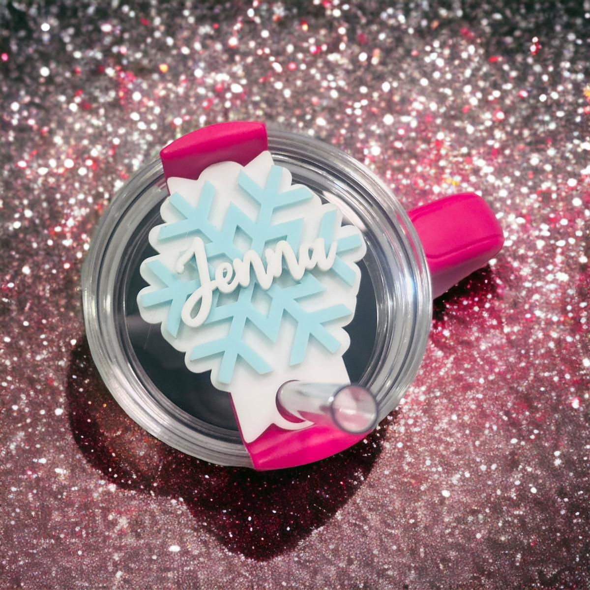 A clear cup with a pink handle and a snowflake-shaped name tag attached to it, placed on a glittery surface.