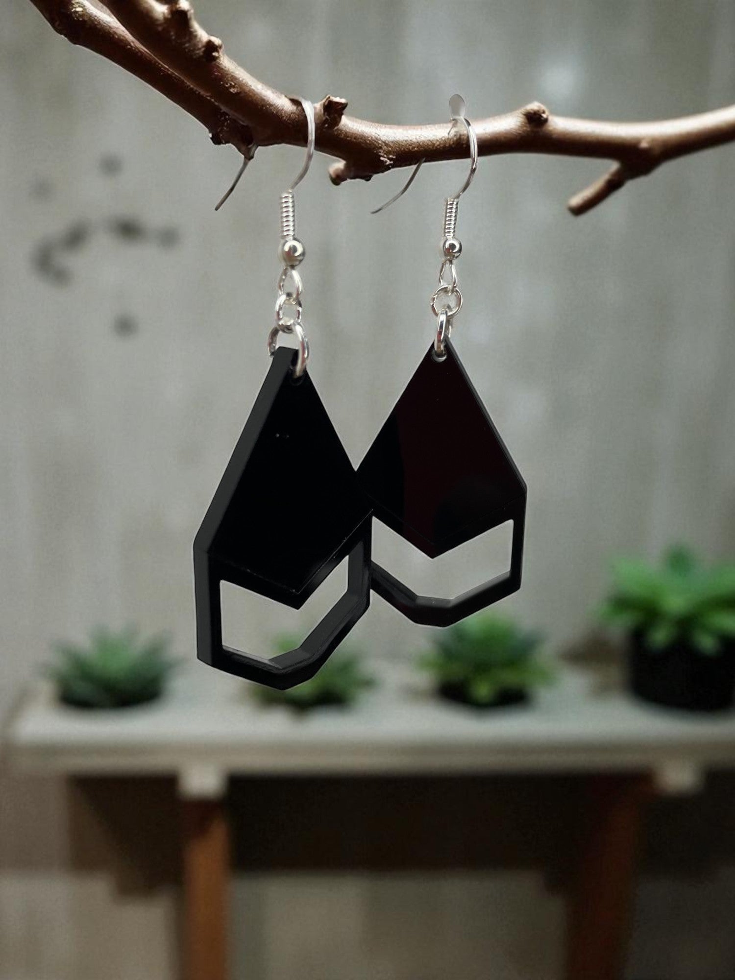 A pair of black geometric earrings hanging from a branch, with a blurred background of plants and a wooden surface.