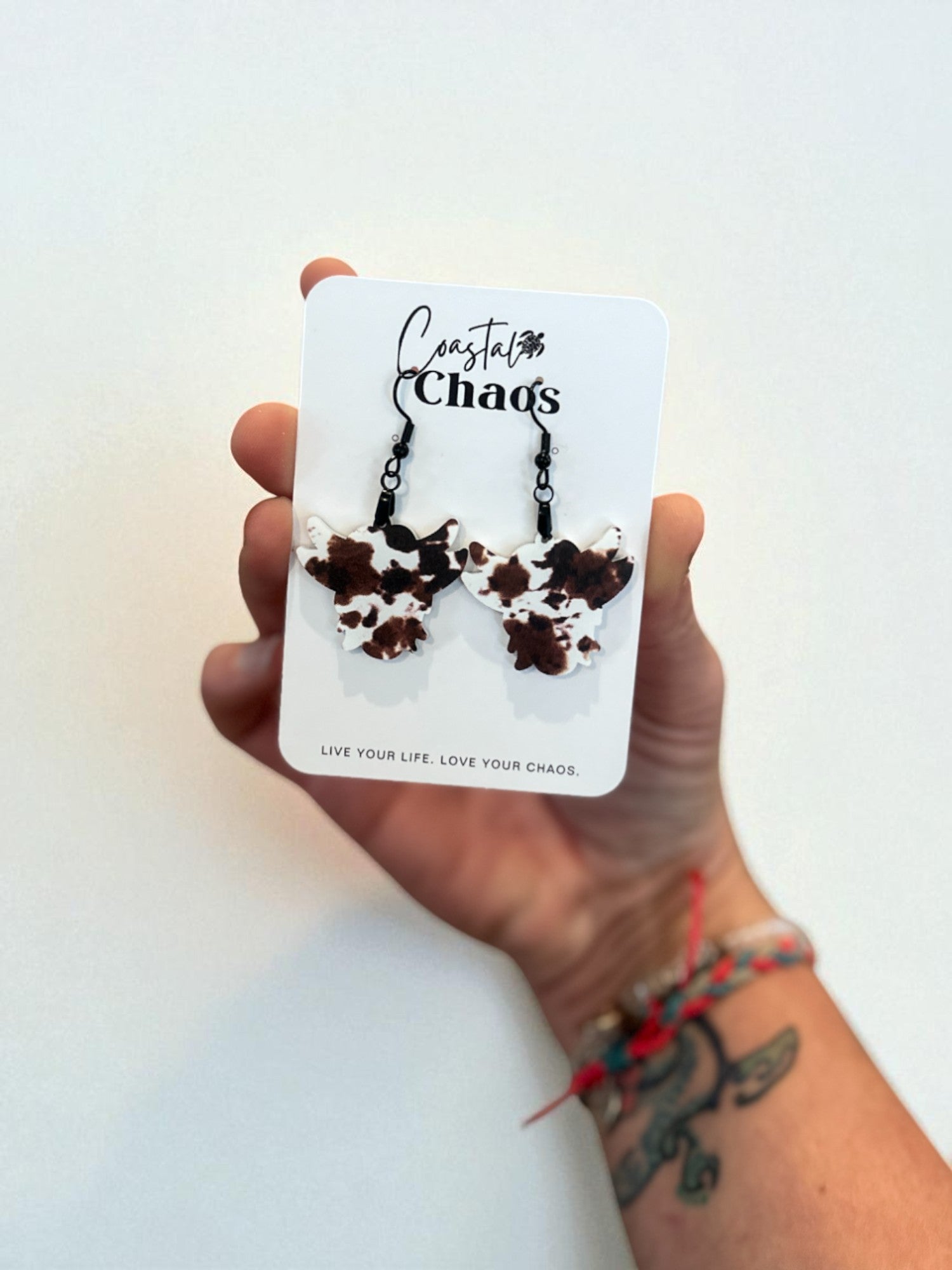 A hand holding a pair of earrings against a white background, with a tattoo visible on the wrist.