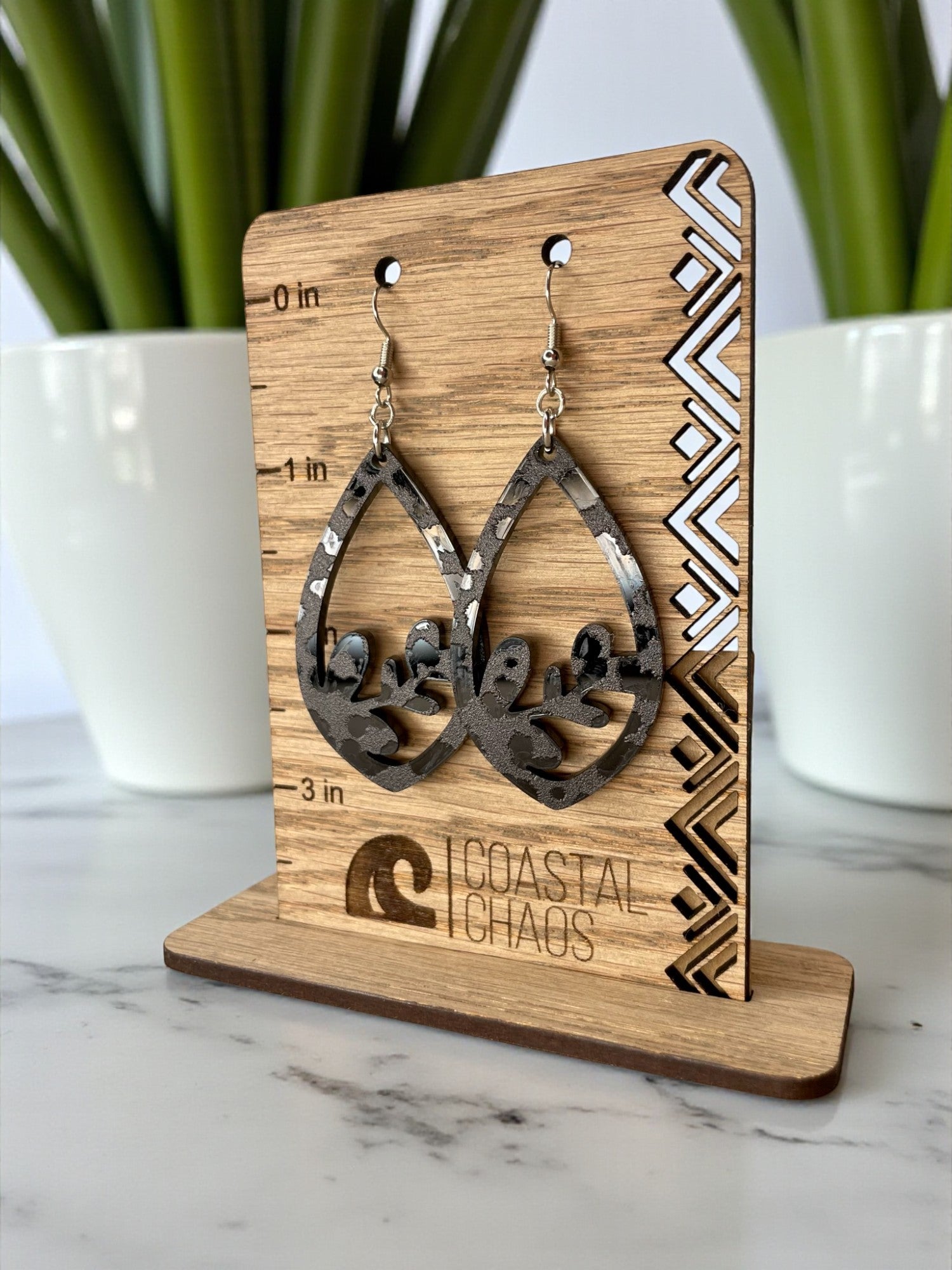 A pair of earrings on a wooden stand, with a decorative pattern on the stand and a plant in the background.