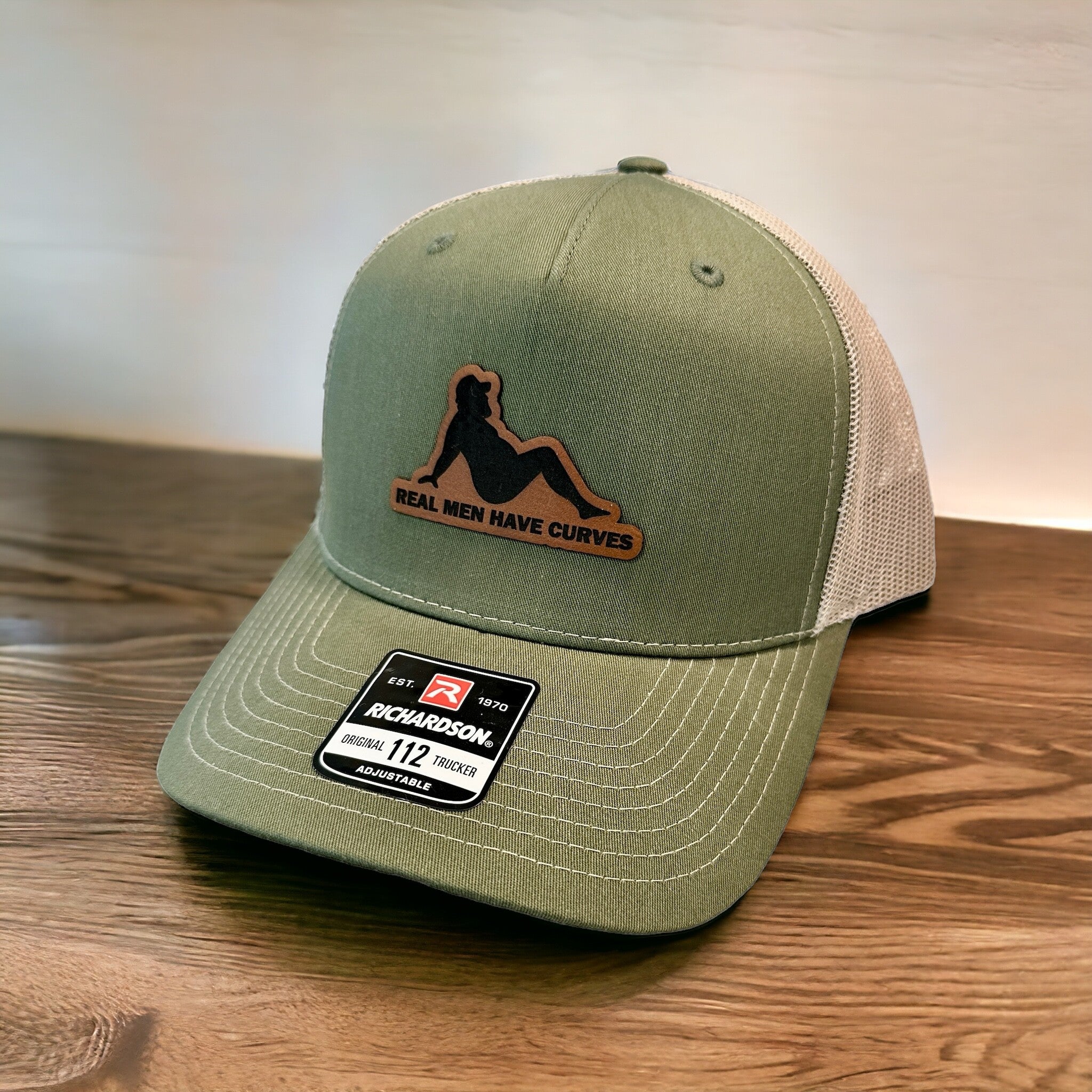 A green baseball cap with a leather patch on the front, displaying the text "Real men have curves" and a silhouette of a woman.