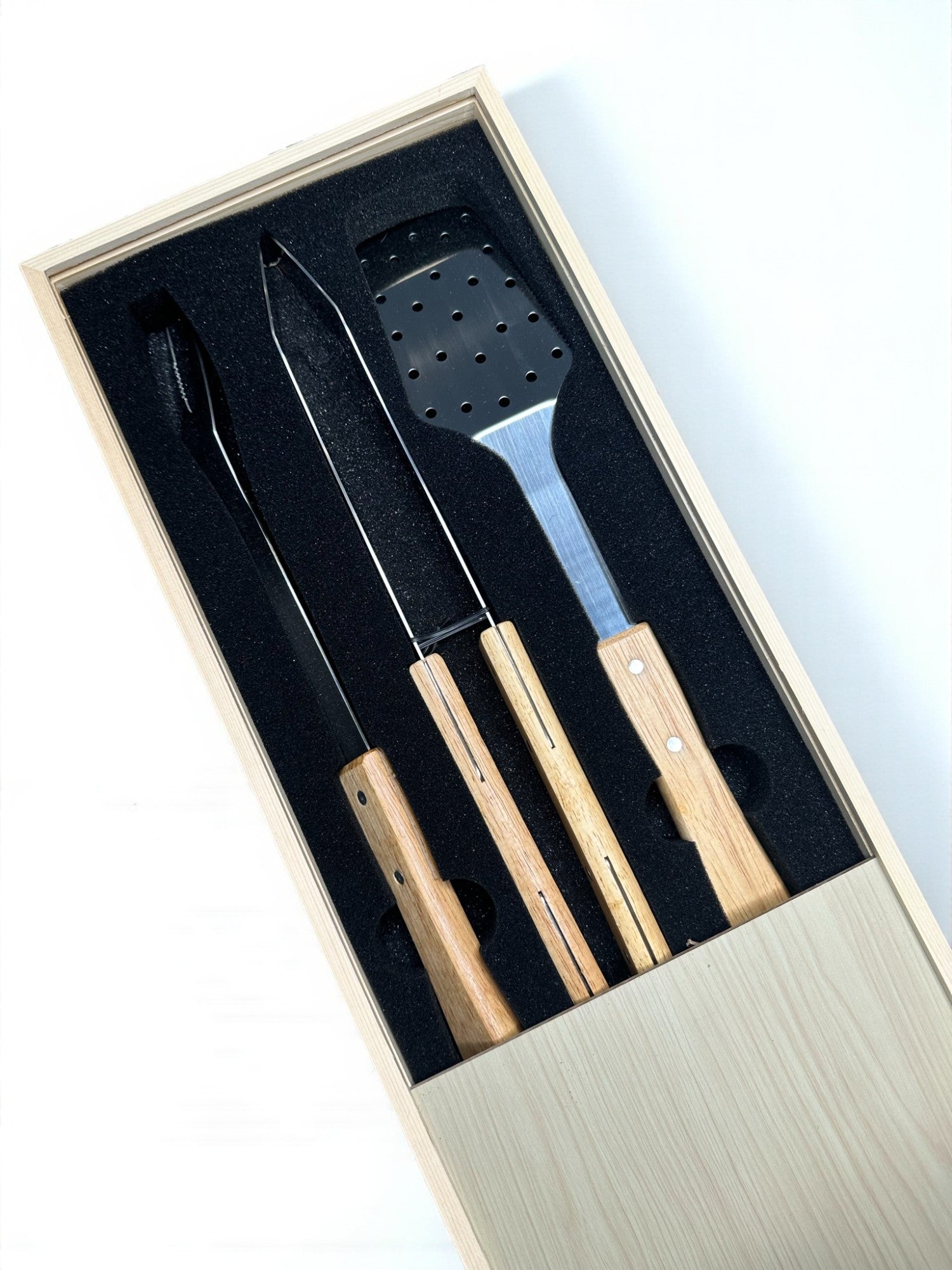 A set of three wooden-handled kitchen utensils, including a spatula and two knives, are neatly arranged in a wooden box with a black interior.
