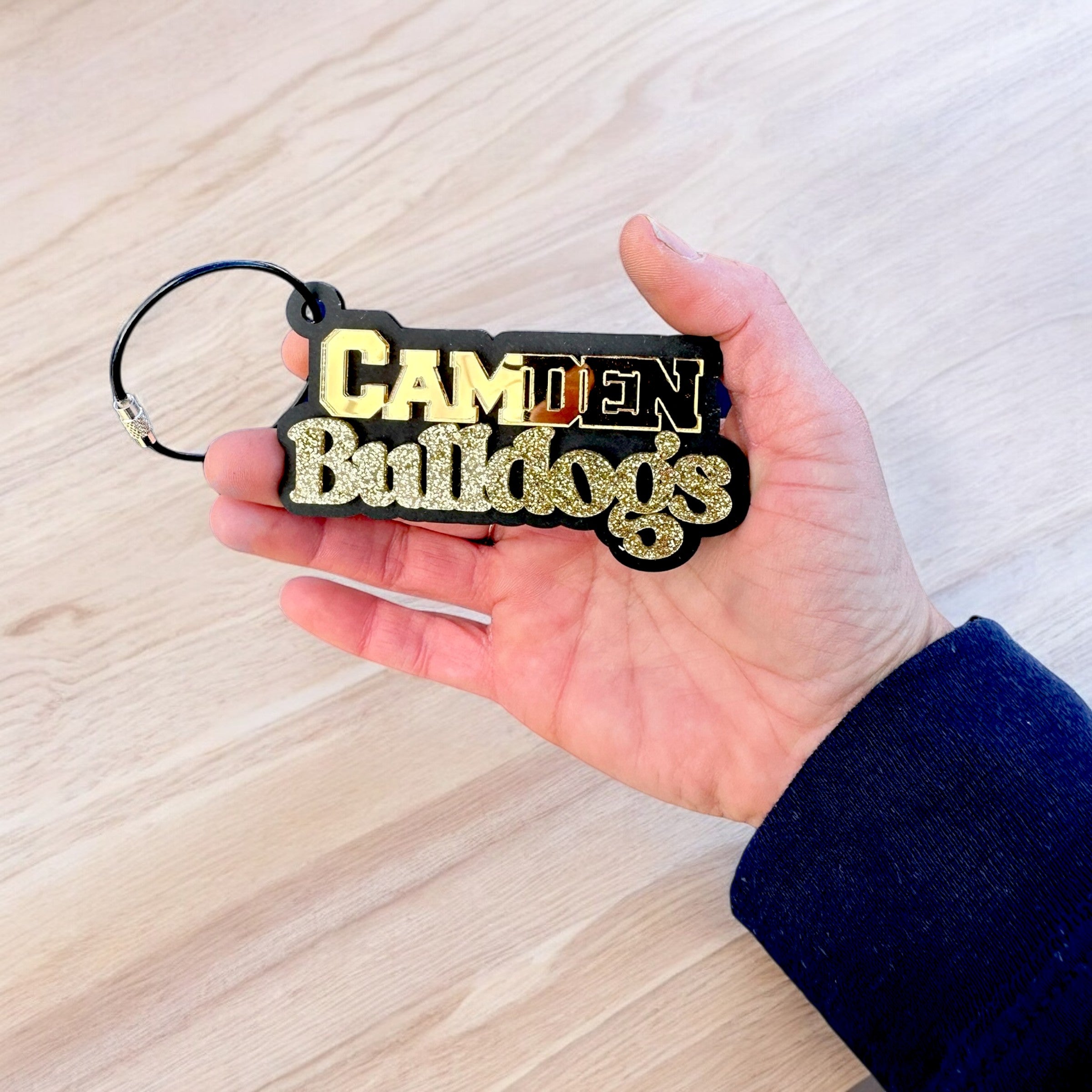 A hand holding a black and gold keychain with the text "Camden Bulldogs" on it.