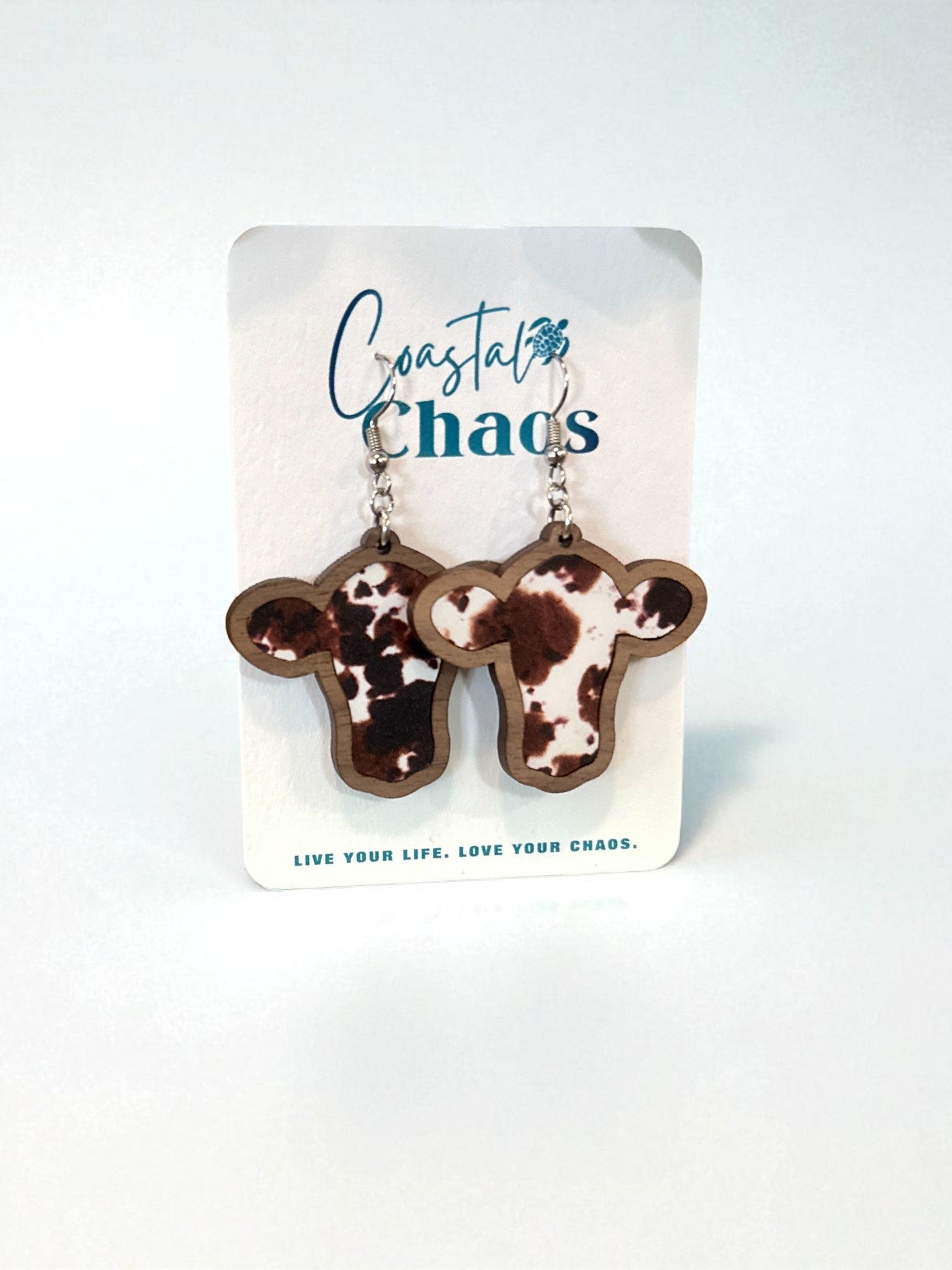 A pair of wooden earrings shaped like cow heads, with the text "Coastal Chaos" and the phrase "Live Your Life. Love Your Chaos" printed on a white background.