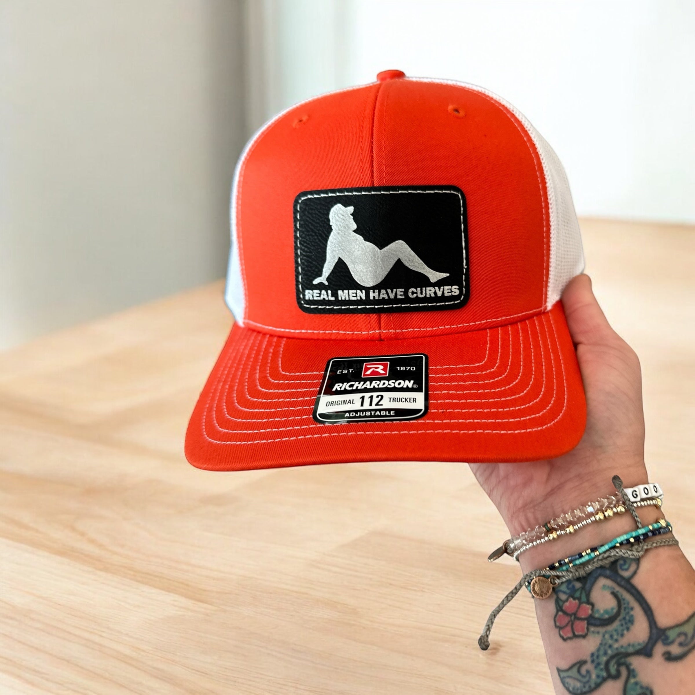 A person is holding up a red baseball cap with a patch on it, which features a graphic of a person in a seated position.