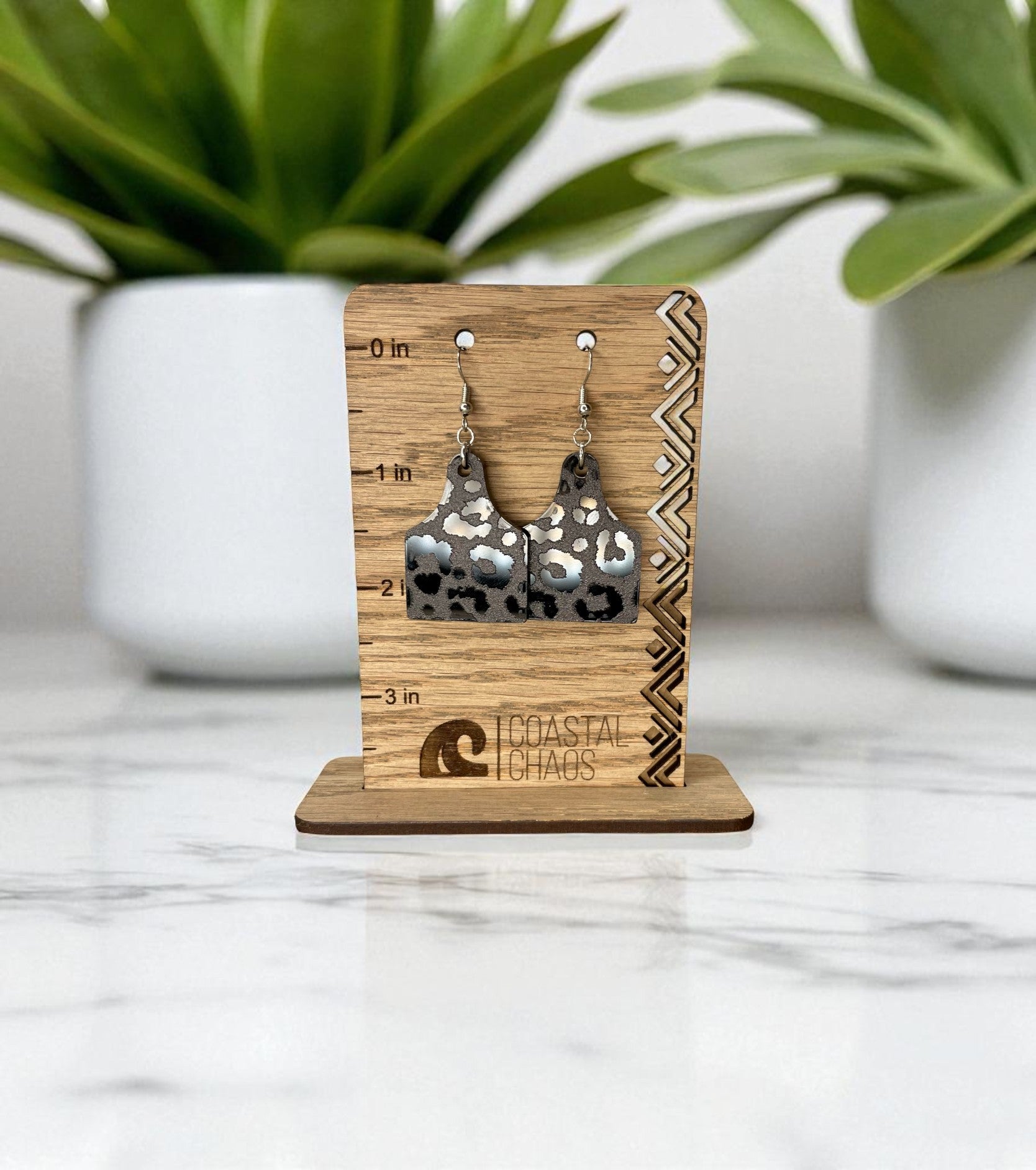 A pair of silver earrings with a leopard print design, displayed on a wooden stand with a ruler for size reference.