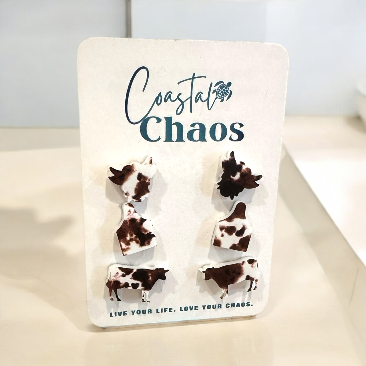a pair of earrings shaped like cows, with the text "Coastal Chaos" and "Live Your Life. Love Your Chaos." displayed on a white background.