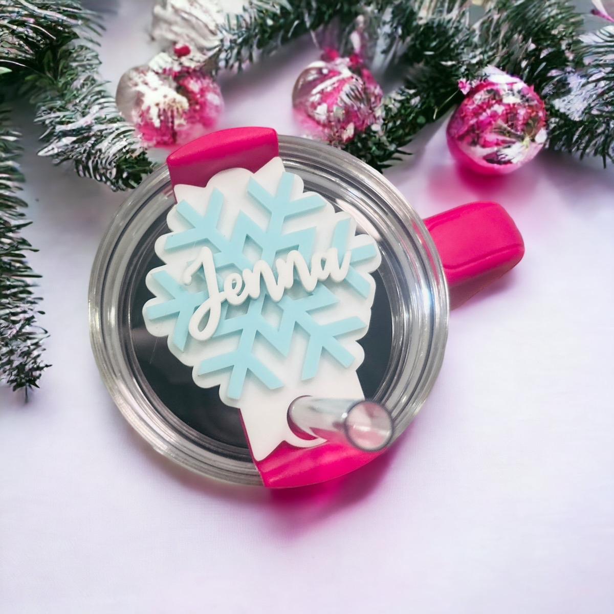 A clear glass cup with a pink handle and a snowflake-shaped lid, decorated with the name "Jenna" written on it. The cup is surrounded by Christmas decorations, including ornaments and pine branches.