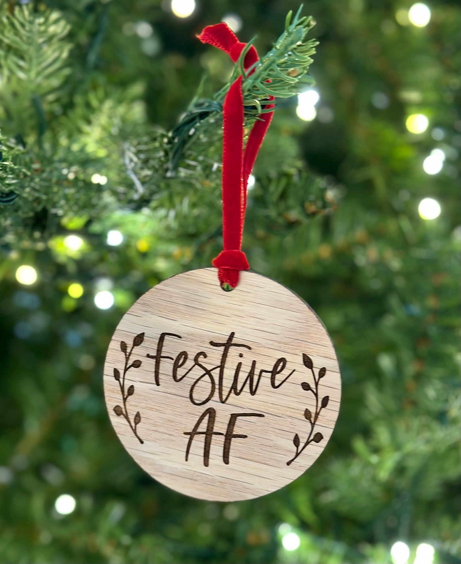 A wooden ornament with the text "Festive AF" engraved on it, hanging from a tree branch with a red ribbon.
