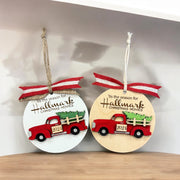 Two circular ornaments hanging on a shelf, each featuring a red truck with a Christmas tree in the bed.