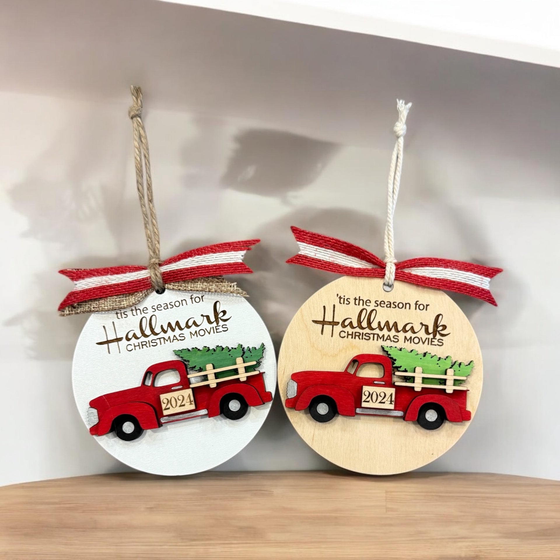 Two circular ornaments hanging on a shelf, each featuring a red truck with a Christmas tree in the bed.