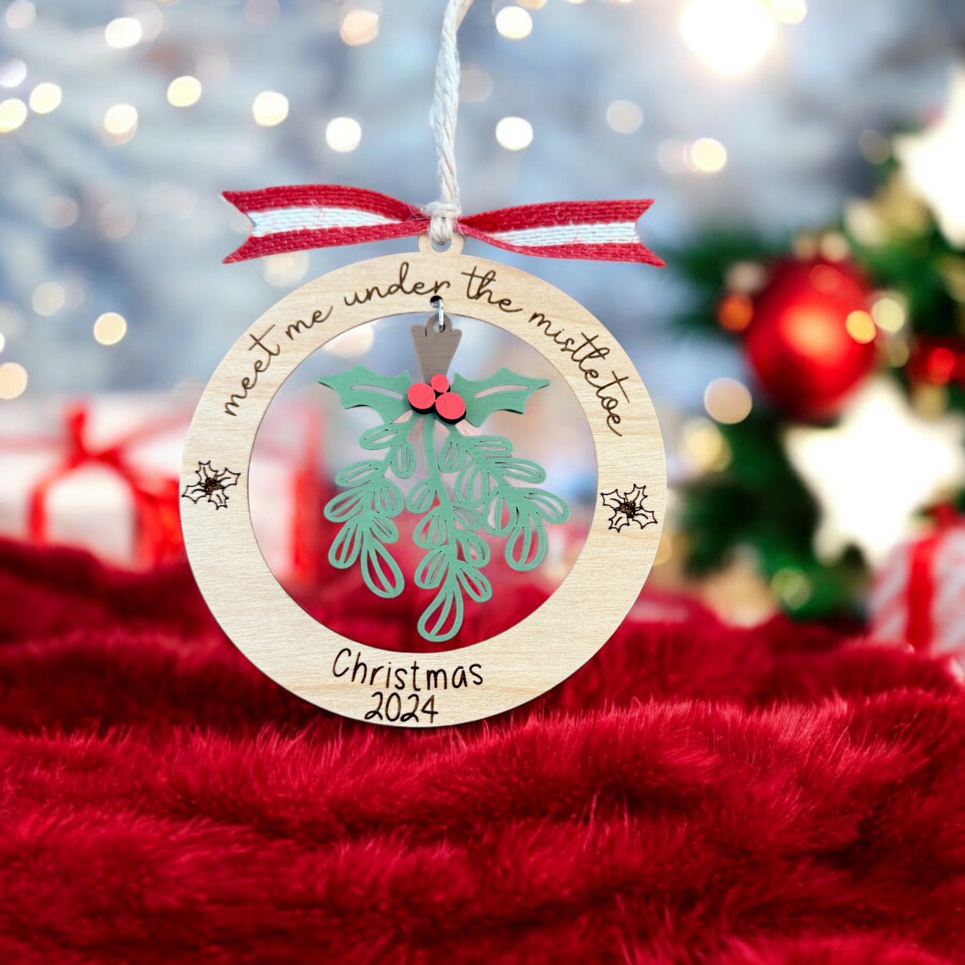 A wooden ornament with a Christmas tree and mistletoe hangs on a string, with a red ribbon and the text "Meet me under the mistletoe" written on it.