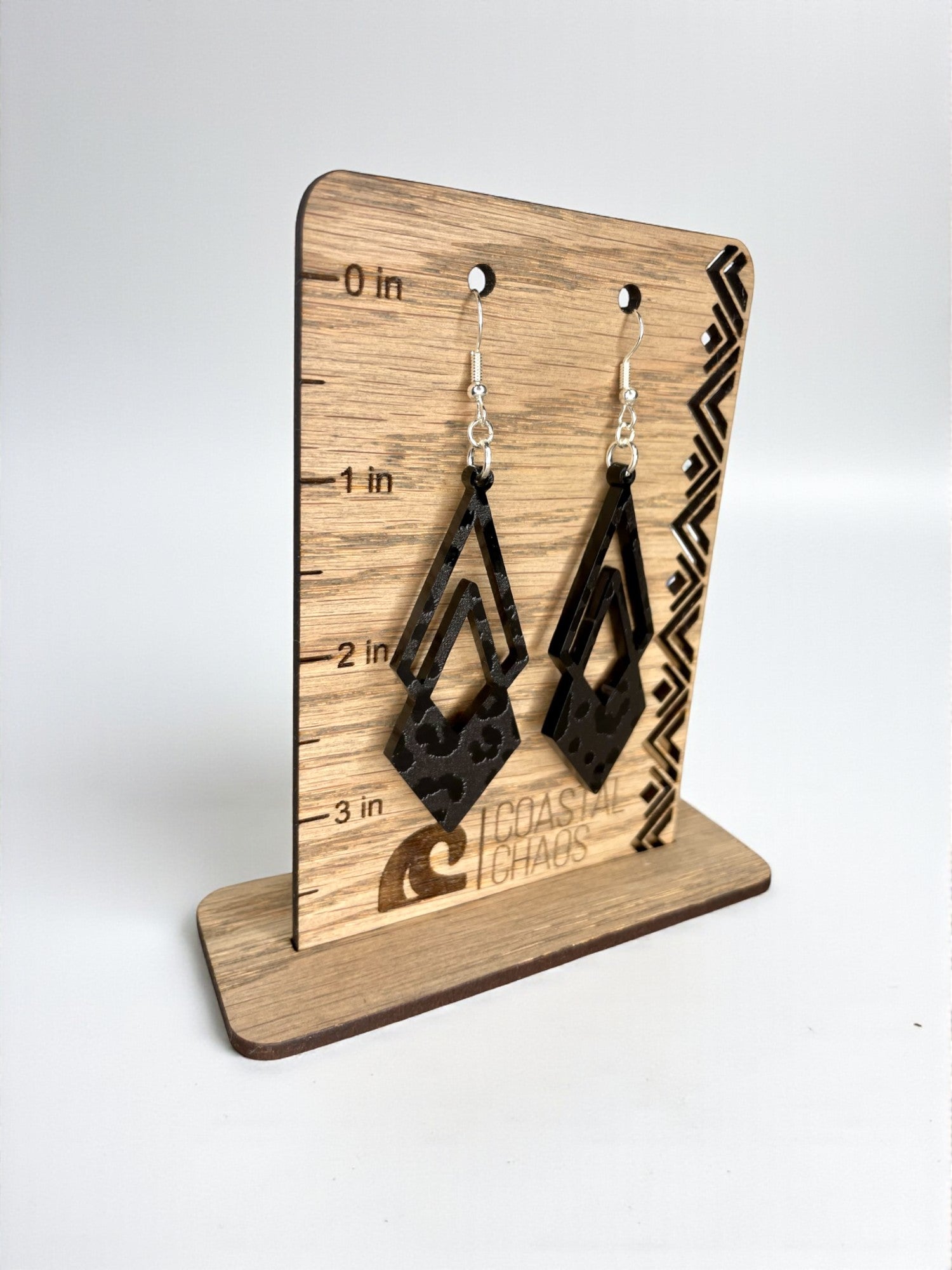 A pair of black earrings with a geometric design, displayed on a wooden stand with measurement markings.