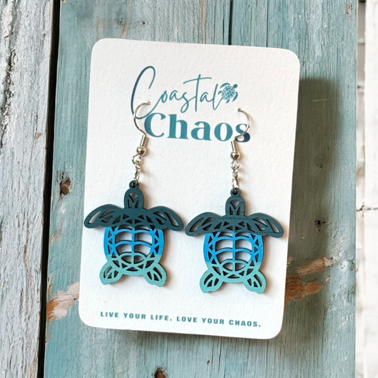 A pair of earrings in the shape of sea turtles, with the earrings hanging from a white card that has the text "Coastal Chaos" and "Live Your Life. Love Your Chaos." printed on it.