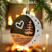 A round ornament hanging on a tree branch, featuring a heart symbol, a crescent moon, and a pine tree design. The ornament has a message that reads "The sky looks different when someone you love is up there".
