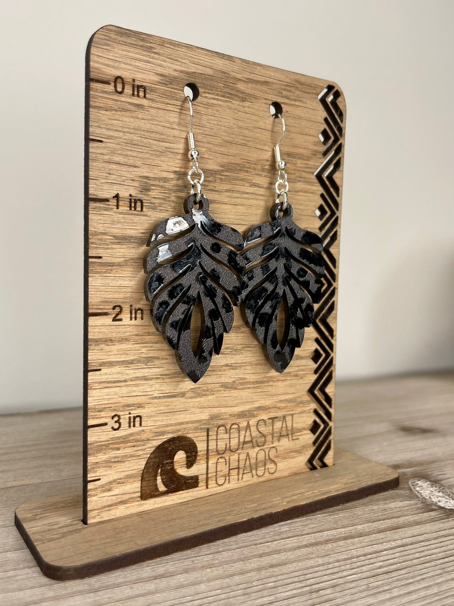 A pair of black leaf-shaped earrings displayed on a wooden stand with measurement markings.