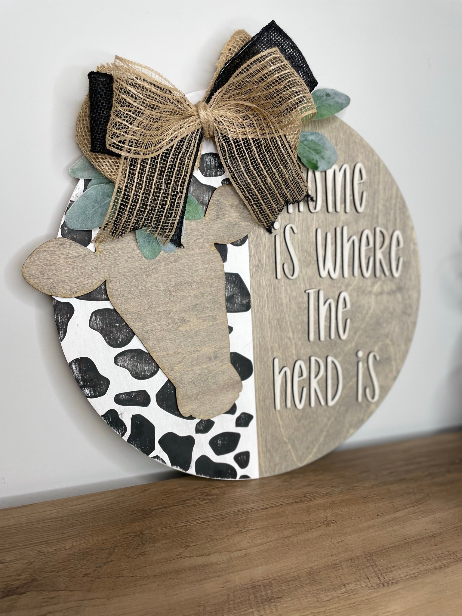 A wooden sign with a cow's head and a decorative bow, along with the text "Home is where the herd is" written on it.