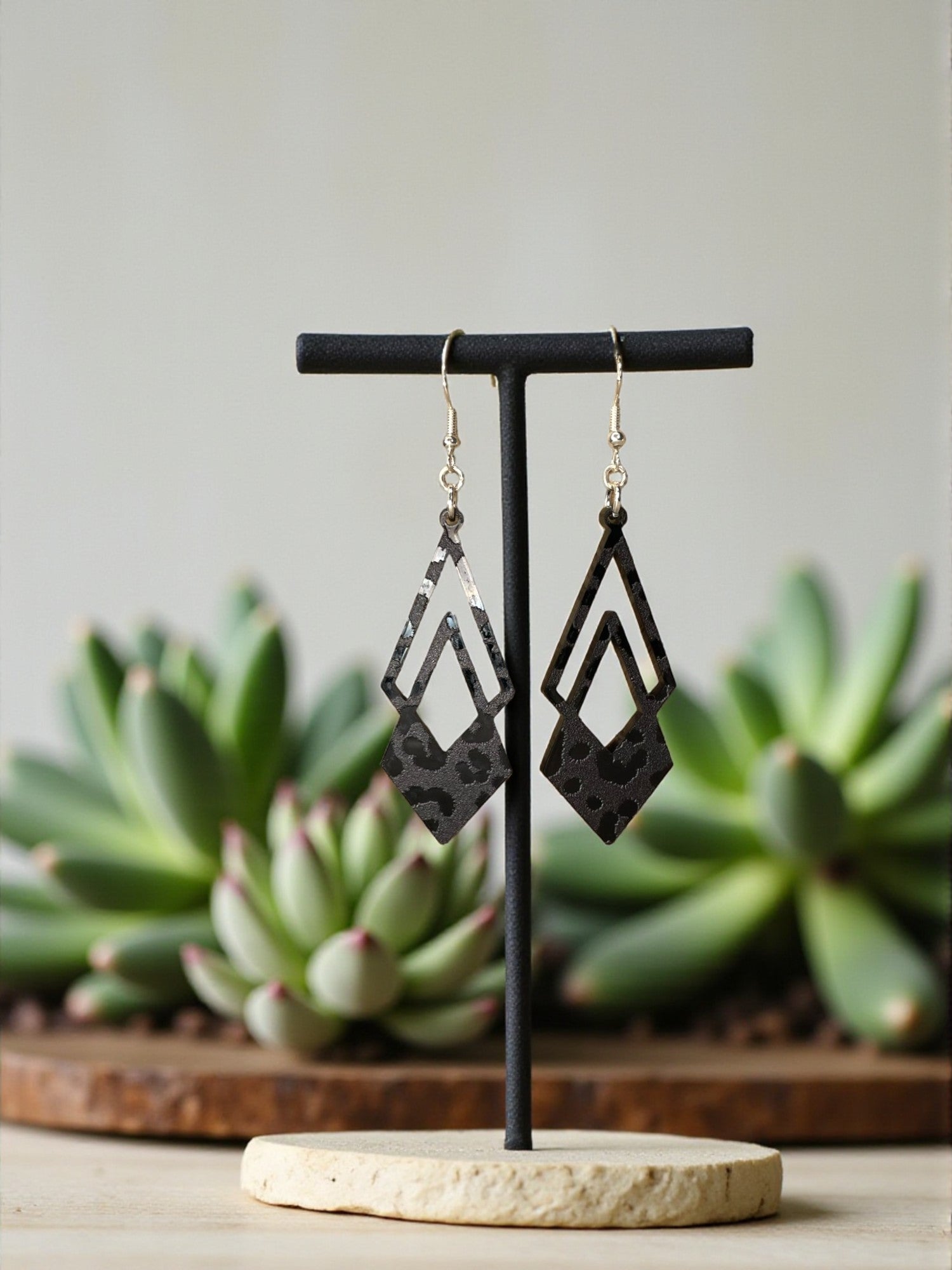 A pair of black geometric earrings with a leopard print design, displayed on a black stand in front of a backdrop of green succulent plants.
