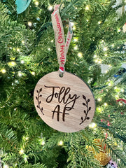 A wooden ornament with the text "Jolly AF" engraved on it, hanging from a Christmas tree.