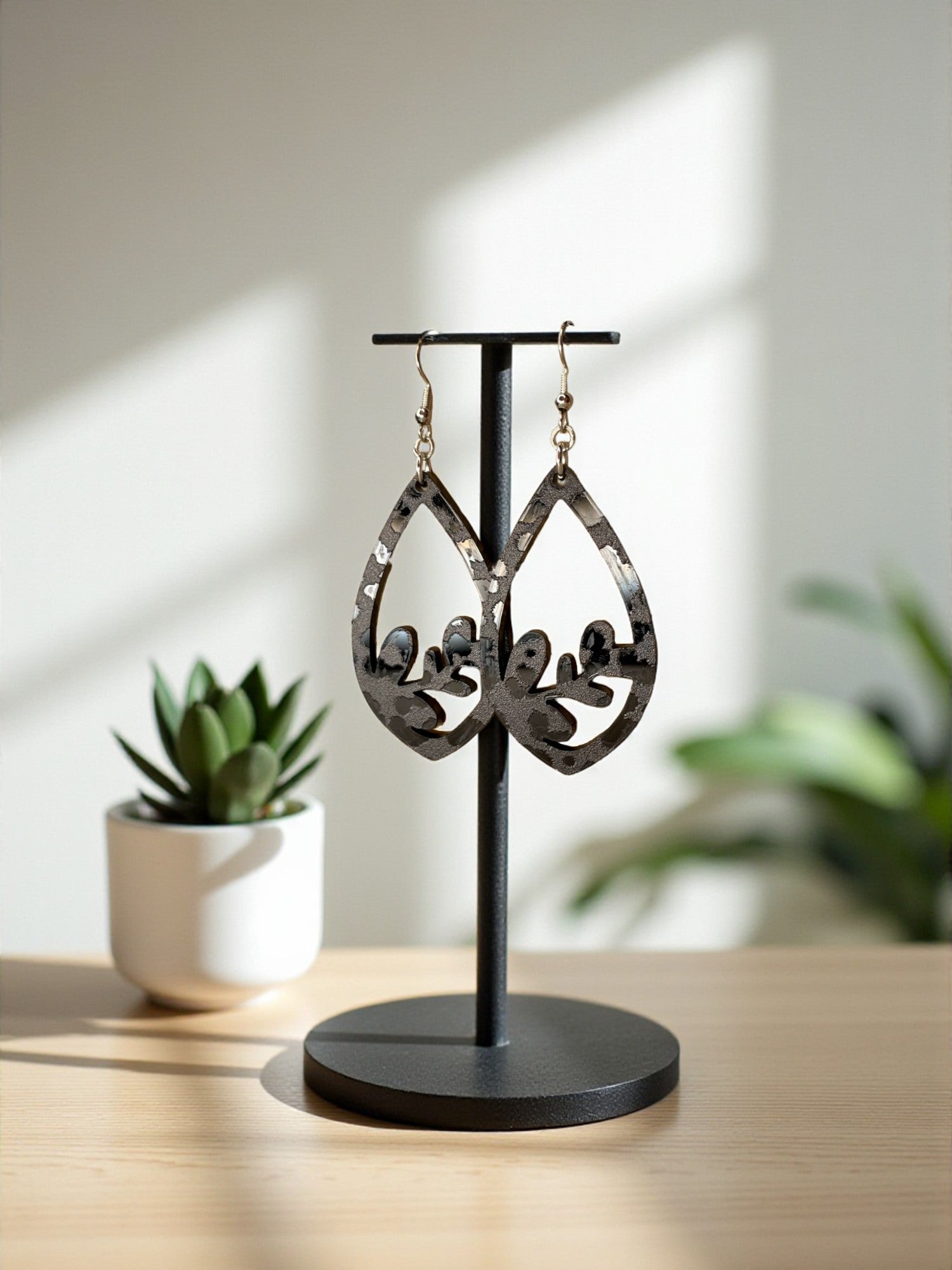 A pair of black and white earrings with a leaf design hangs on a black stand, placed on a wooden surface next to a white pot with a green plant.