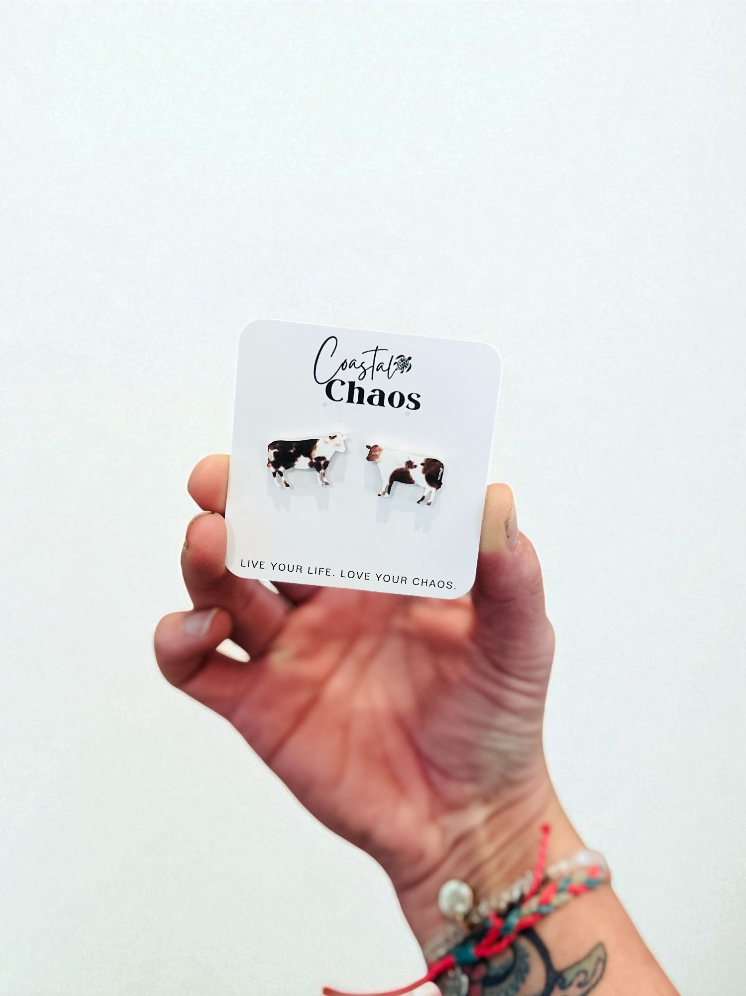 A hand holding a small white card with a pair of cow earrings on it, against a plain white background.