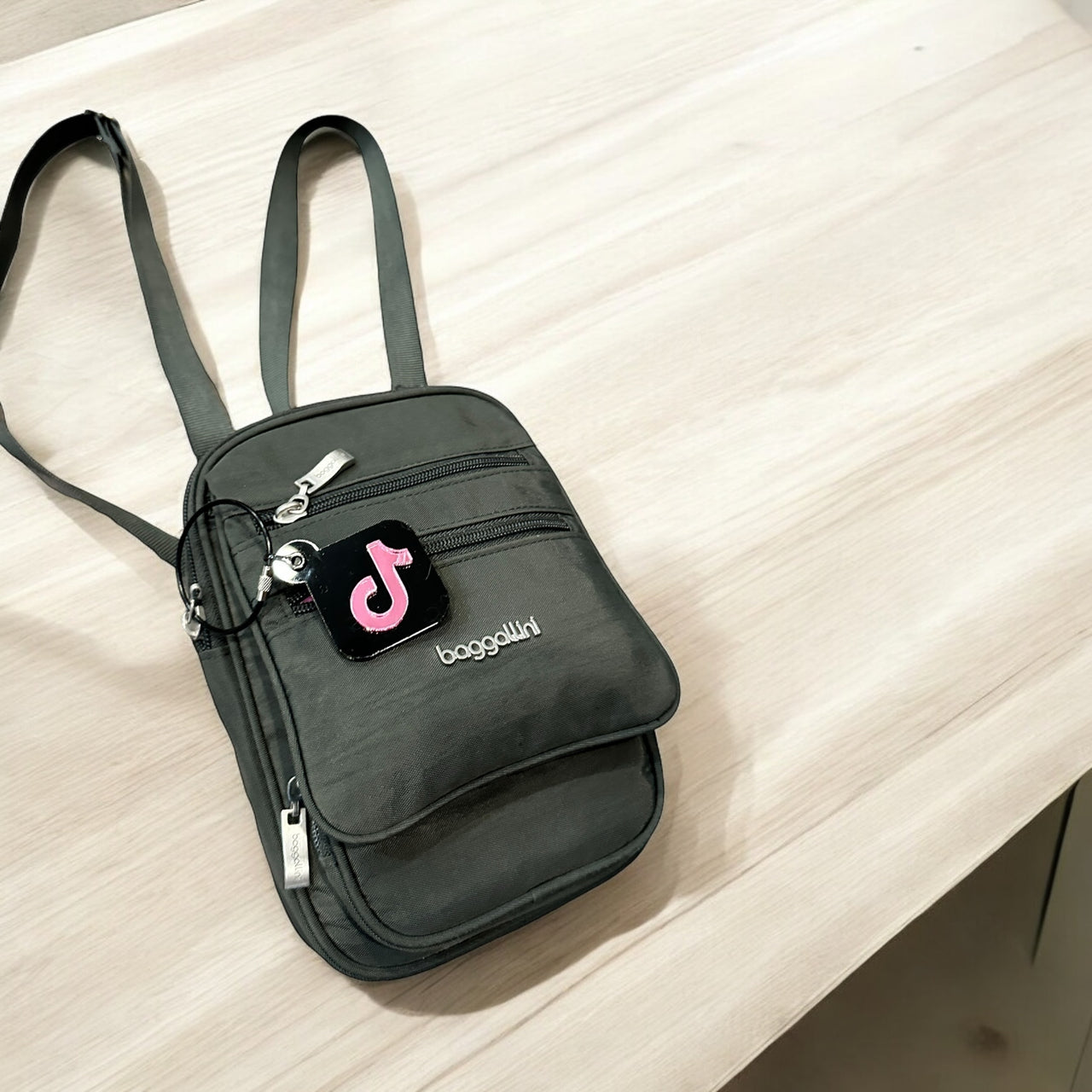 A black crossbody bag with a pink Swoosh logo and the text "baggallini" printed on it, placed on a wooden surface.