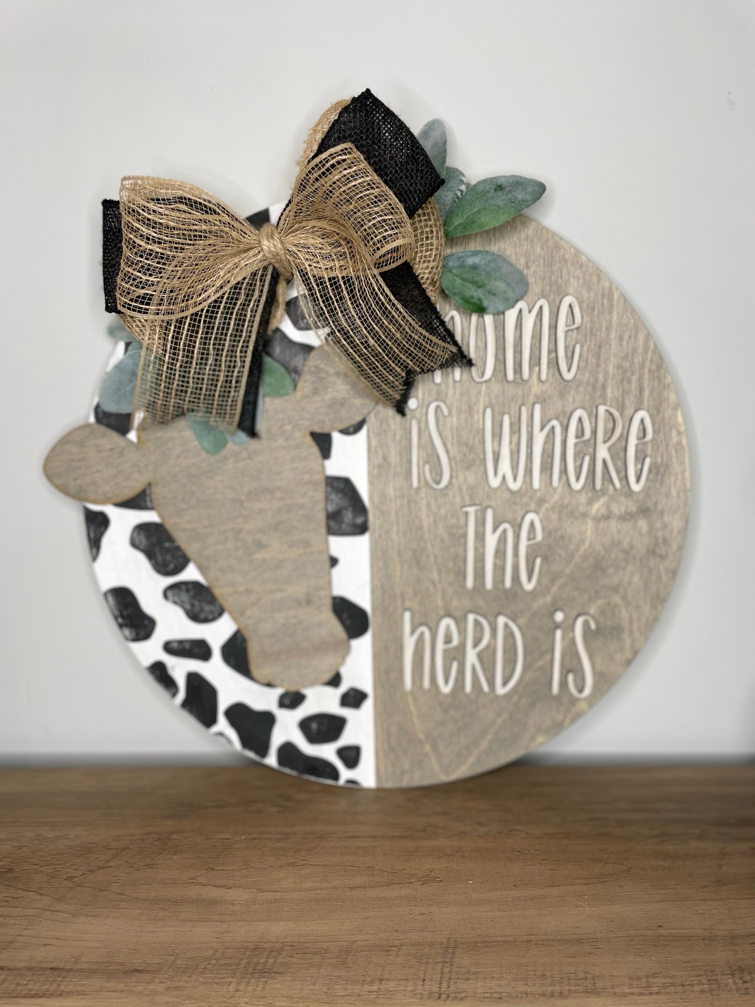 A wooden sign with a cow's head and a decorative bow, along with the text "Home is where the herd is" written on it.