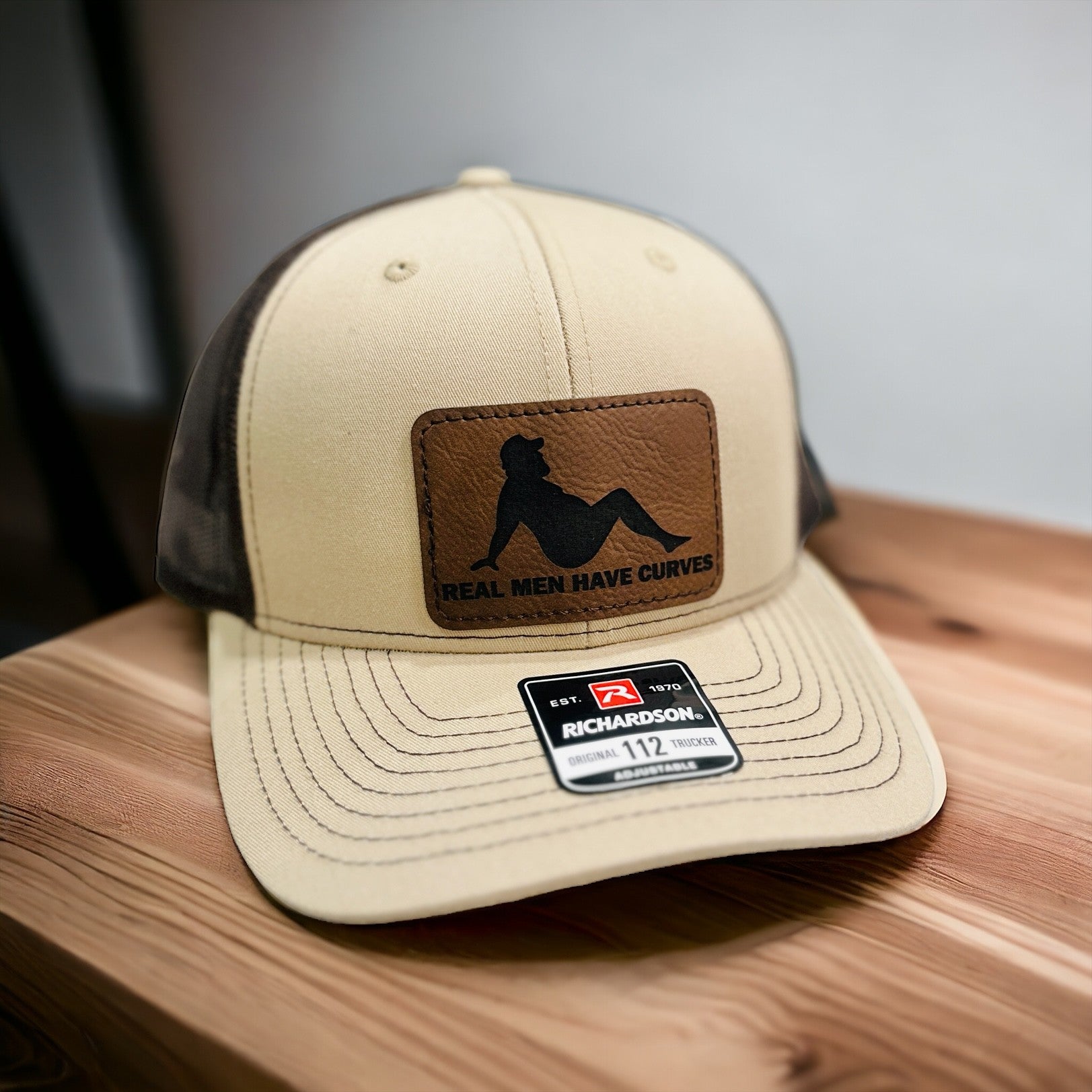 A tan baseball cap with a brown leather patch on the front, displaying the text "Real Men Have Curves" and the brand name "Richardson 112".
