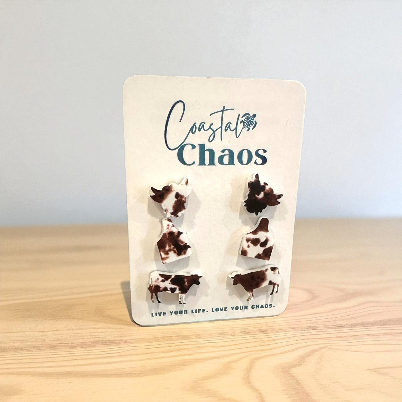 A pair of earrings in the shape of cows, with the text "Coastal Chaos" and "Live Your Life, Love Your Chaos" printed on a white background.
