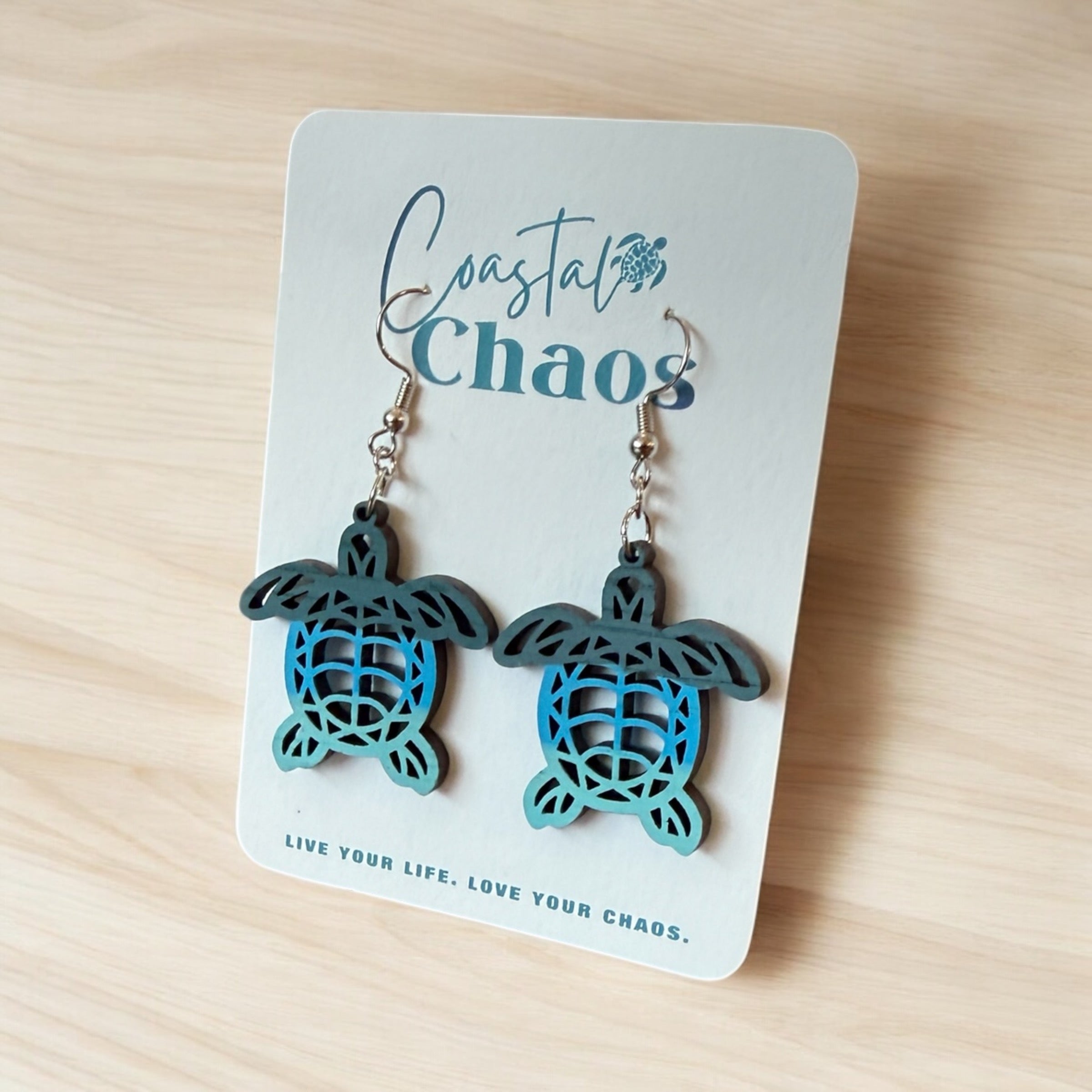 A pair of earrings in the shape of a turtle, displayed on a card with the text "Coastal Chaos" and a motivational quote.