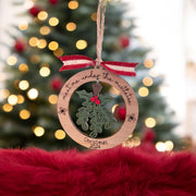 A Christmas ornament with a holly leaf and mistletoe design, hanging from a string with a red ribbon. The ornament is placed on a red fur surface, with a blurred Christmas tree in the background.
