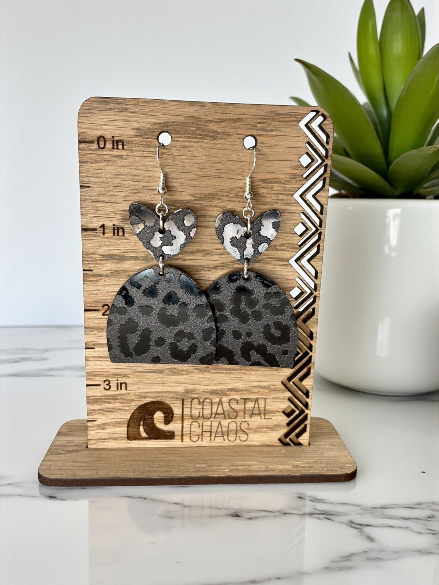 A pair of leopard print earrings displayed on a wooden stand, with a potted plant in the background.