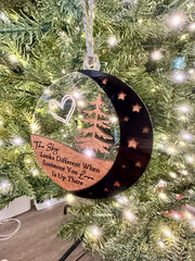 A clear ornament with a crescent moon and stars hanging from a Christmas tree.