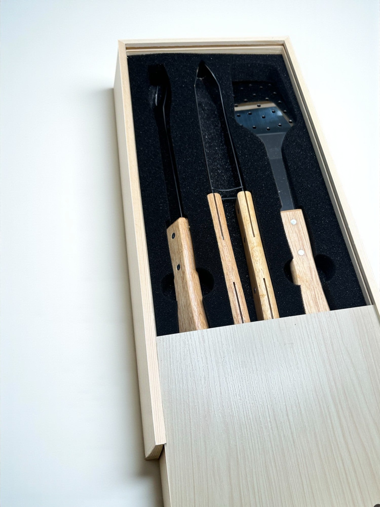 A wooden box containing a set of three wooden utensils, including a spatula and a pair of tongs, placed inside a black foam padding.
