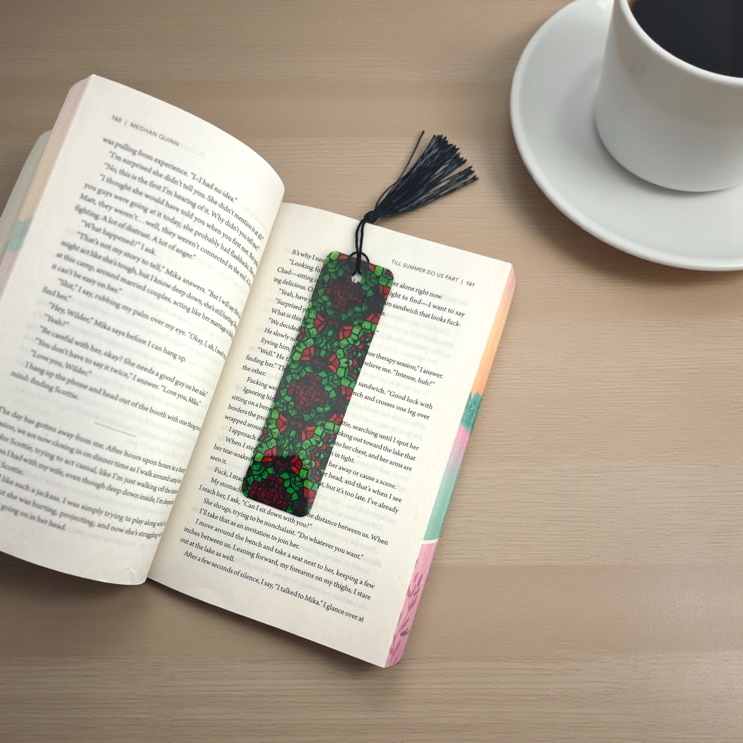 An open book with a floral patterned bookmark is placed on a wooden surface, accompanied by a white coffee cup and a saucer.