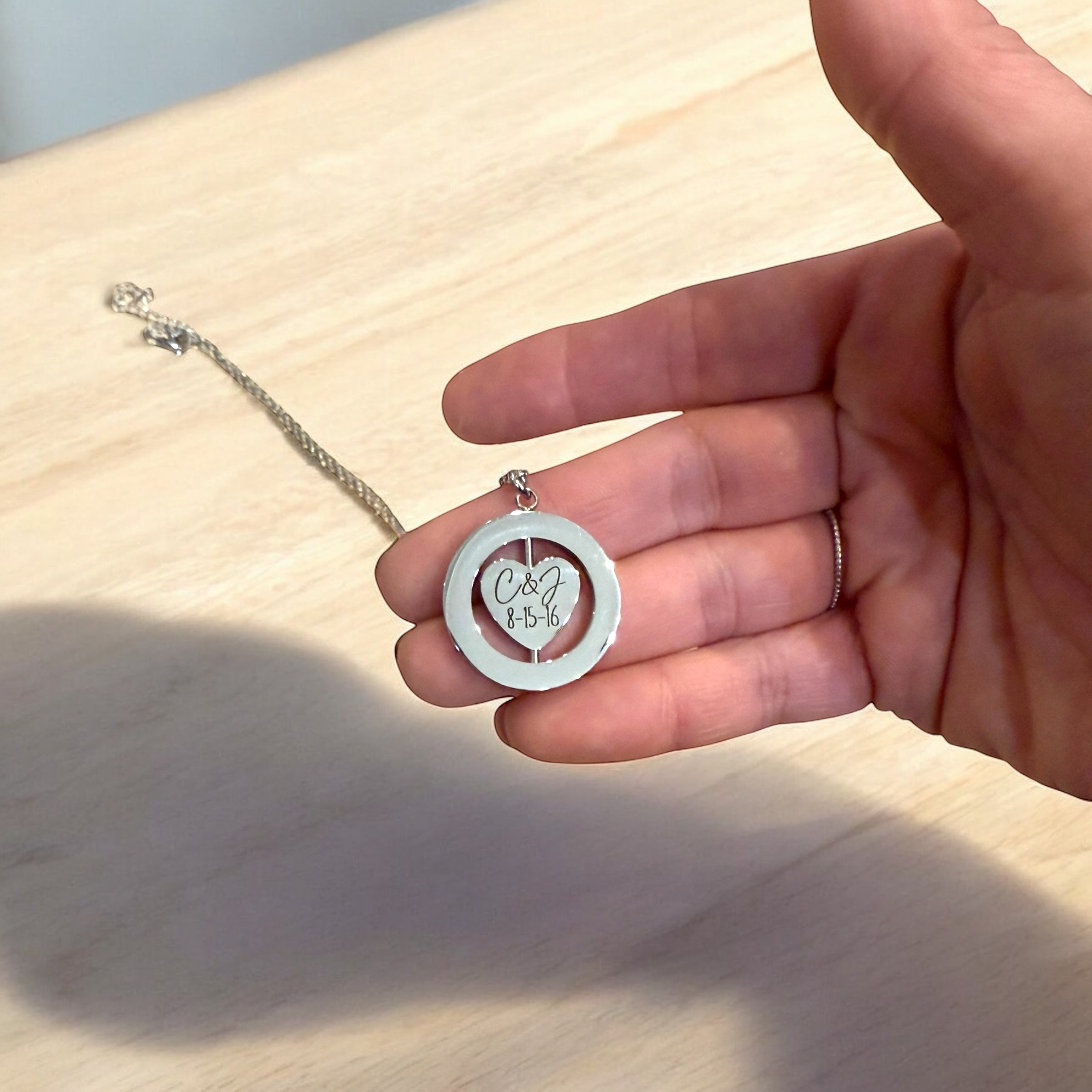 Hand holding a silver necklace with a circular pendant featuring initials and date on a wooden surface.