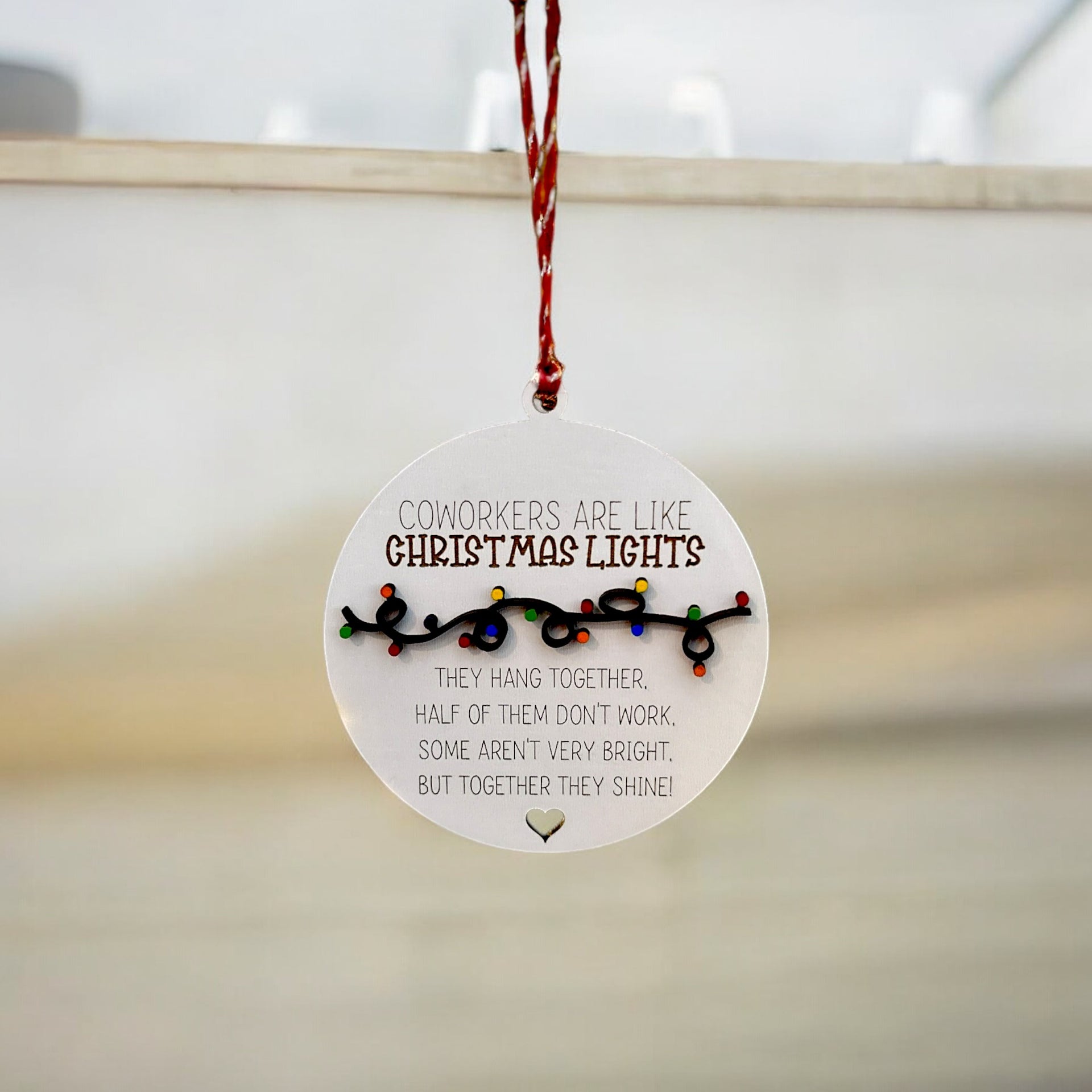 A round ornament hanging from a string, featuring a humorous Christmas-themed message about coworkers.