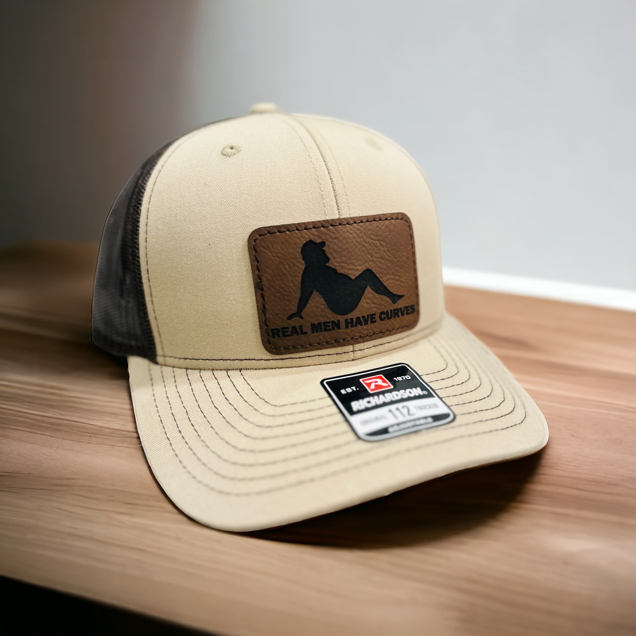 A tan baseball cap with a black patch on the front, displaying the text "Real men have curves" and a silhouette of a person.