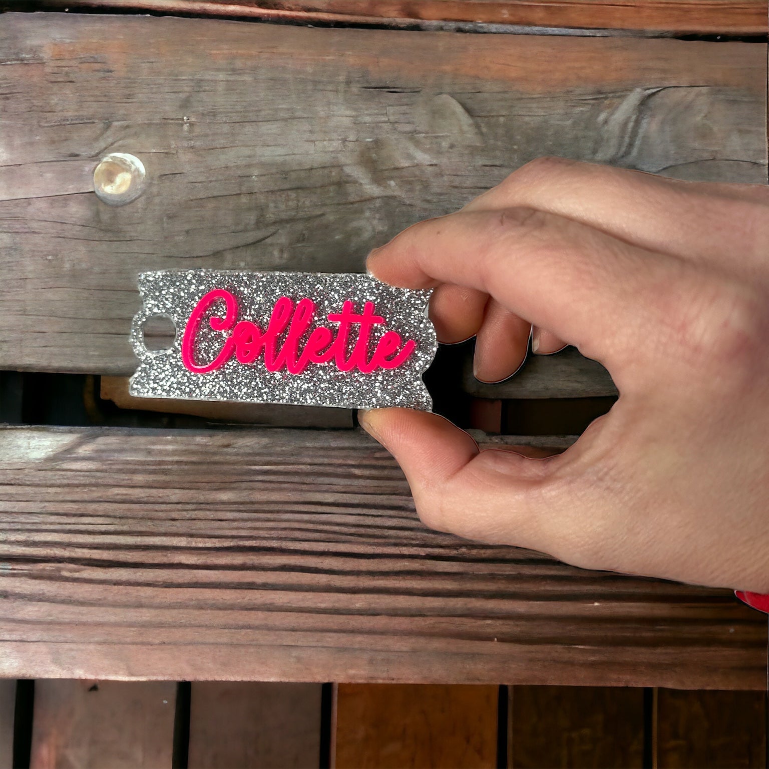 A hand is holding a silver and pink name tag with the name "Colllette" written on it.