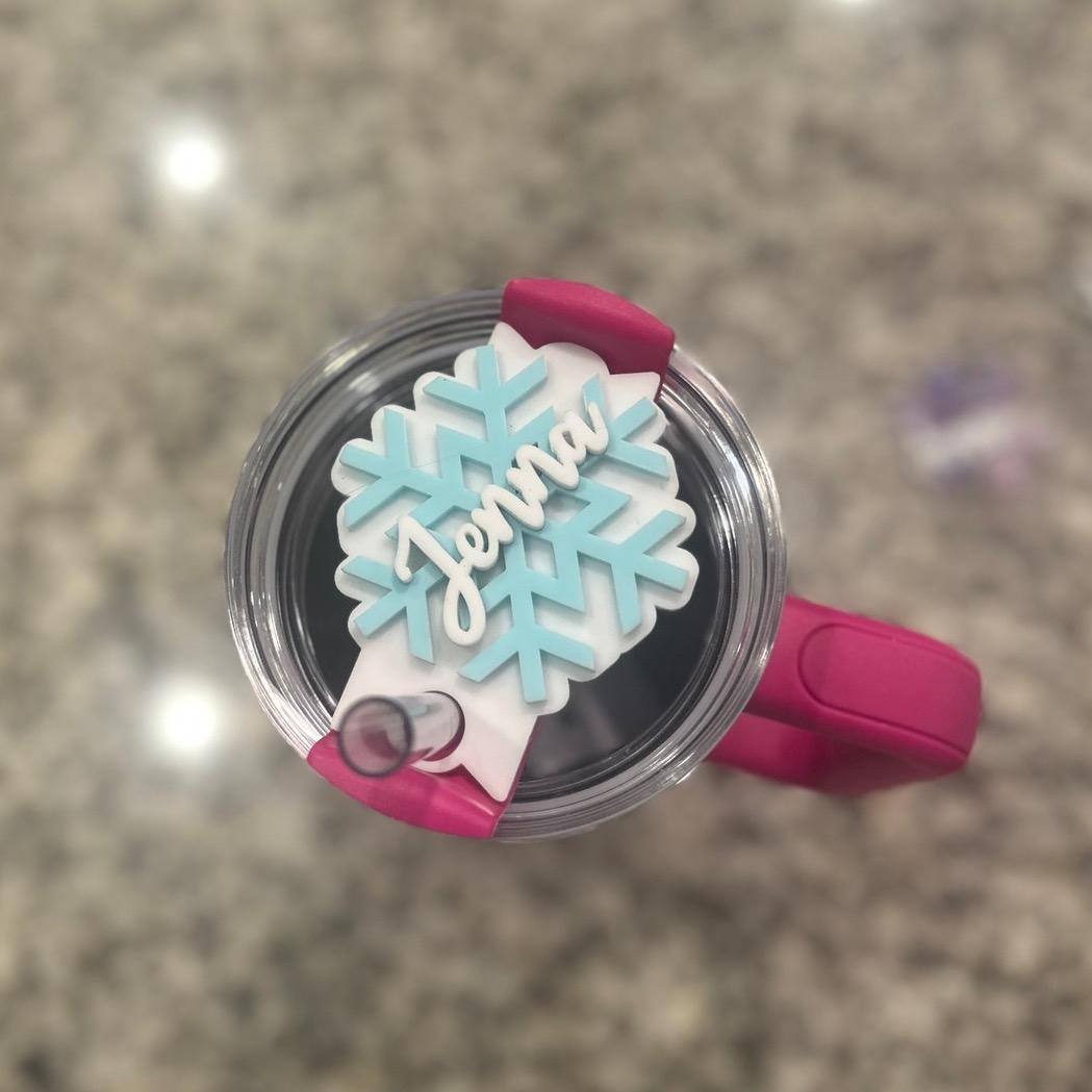 A pink and white snowflake-shaped lid with the name "Jenna" written on it, attached to a clear plastic cup.