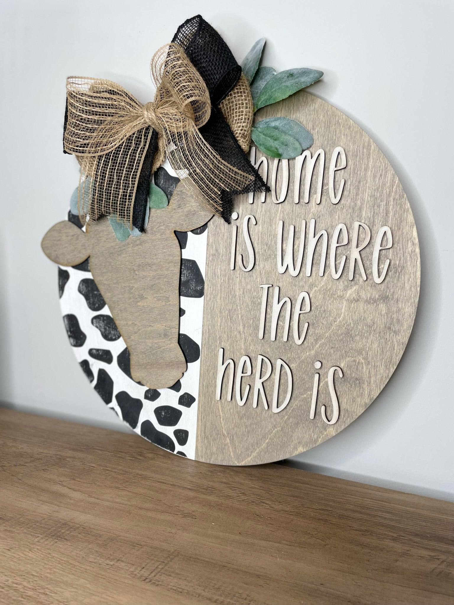 A wooden sign with the text "Home is where the herd is" and a decorative cow silhouette, placed on a wooden surface.