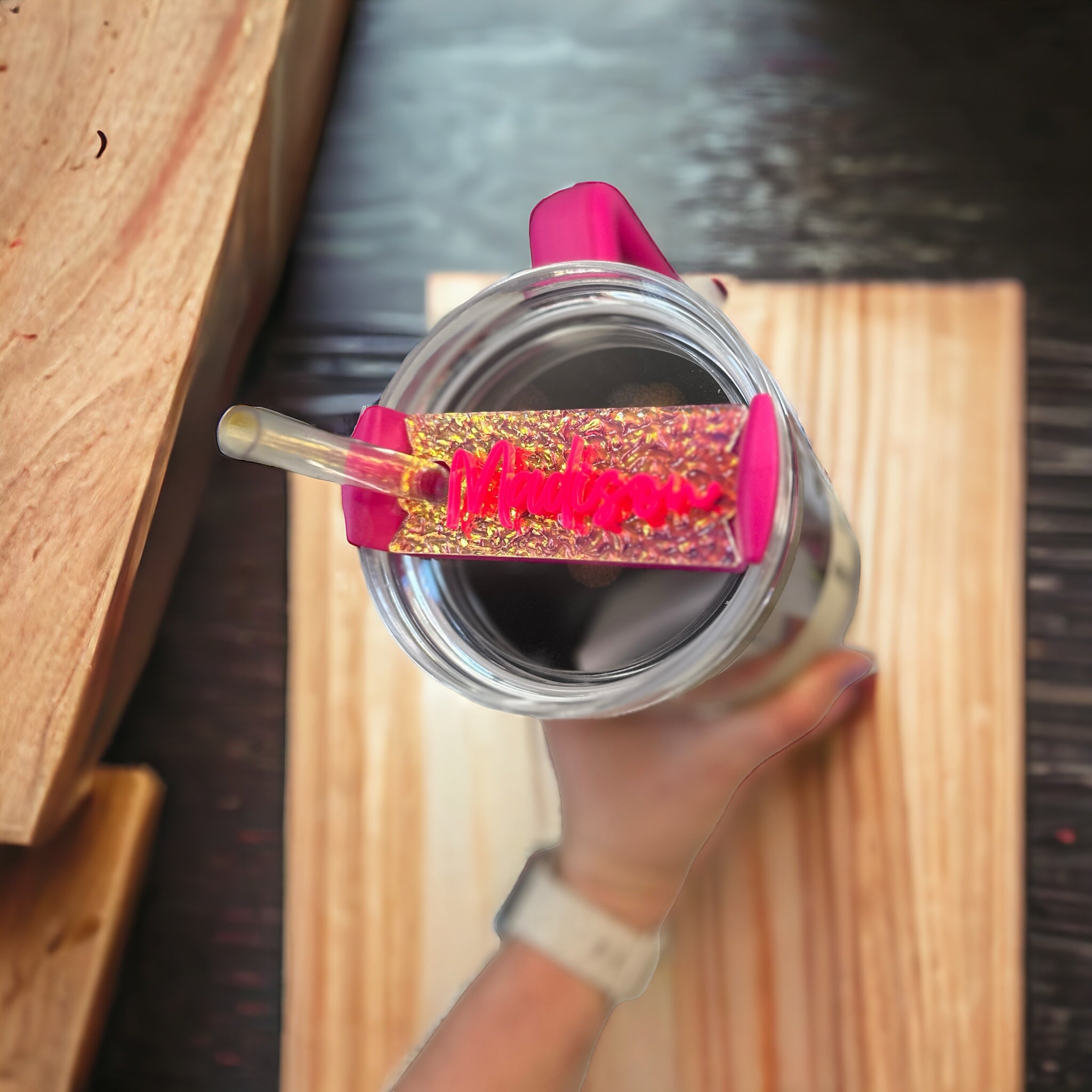 A person's hand holding a clear glass with a pink and gold glittery straw attached to it, placed on a wooden surface.