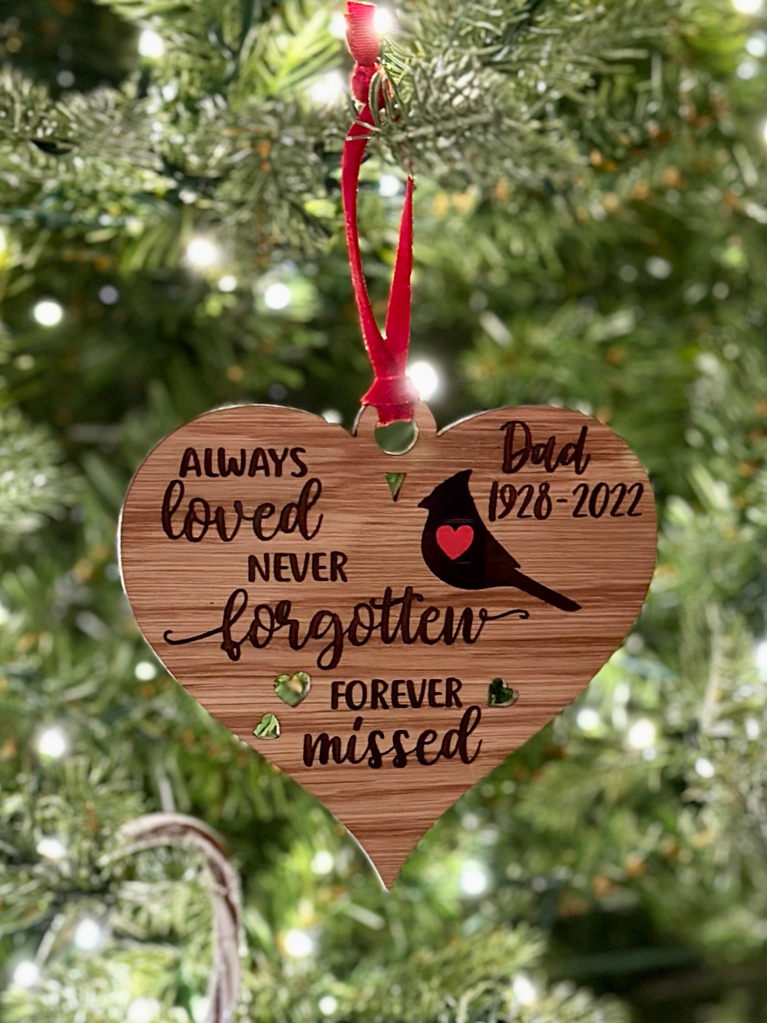A wooden ornament hanging on a Christmas tree, with a heart-shaped design and a message that says "Always loved never forgotten forever missed" and a bird silhouette.