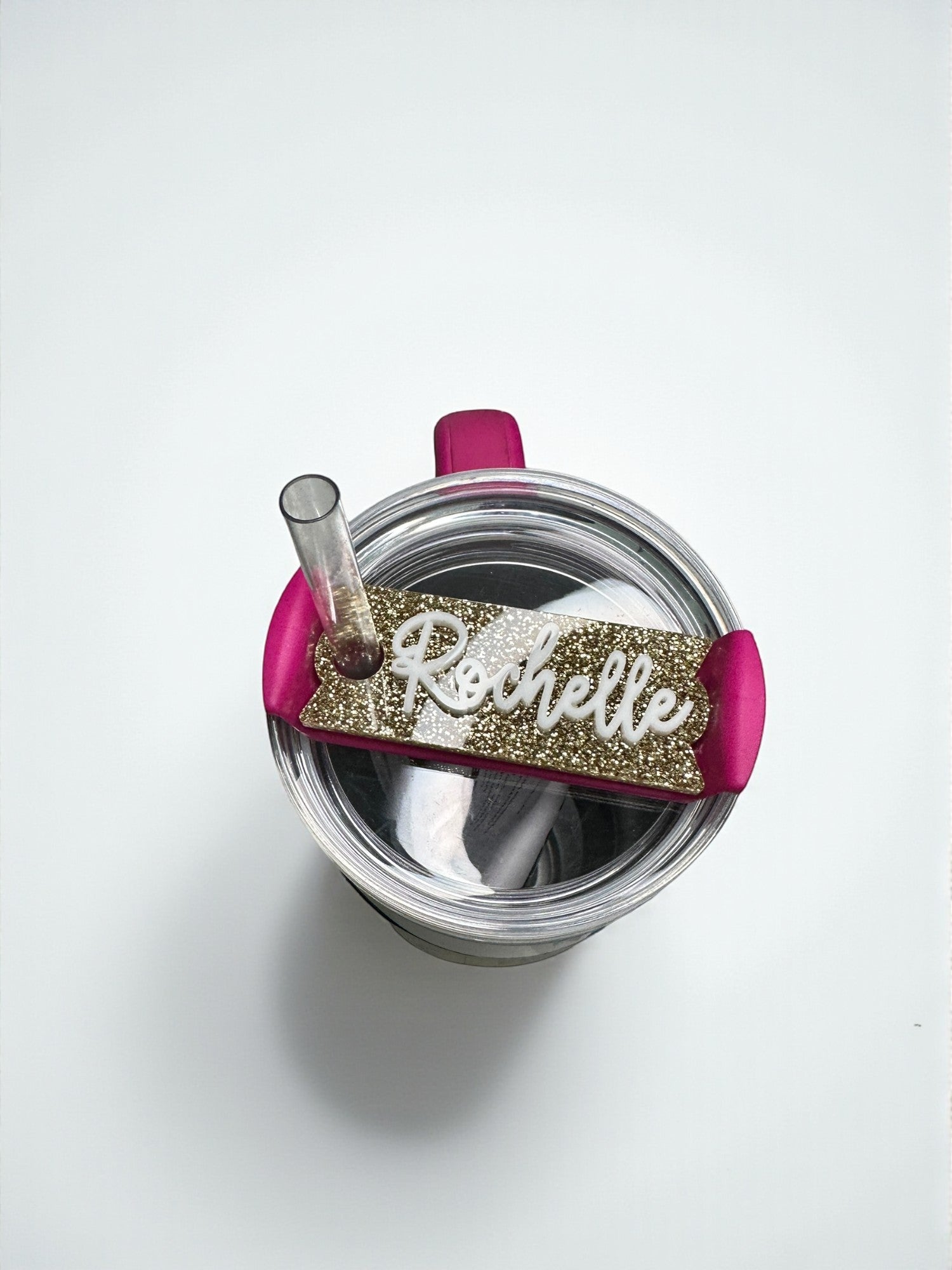 A pink and gold personalized name tag is attached to the lid of a clear glass container, which appears to be a tumbler or a small cup.
