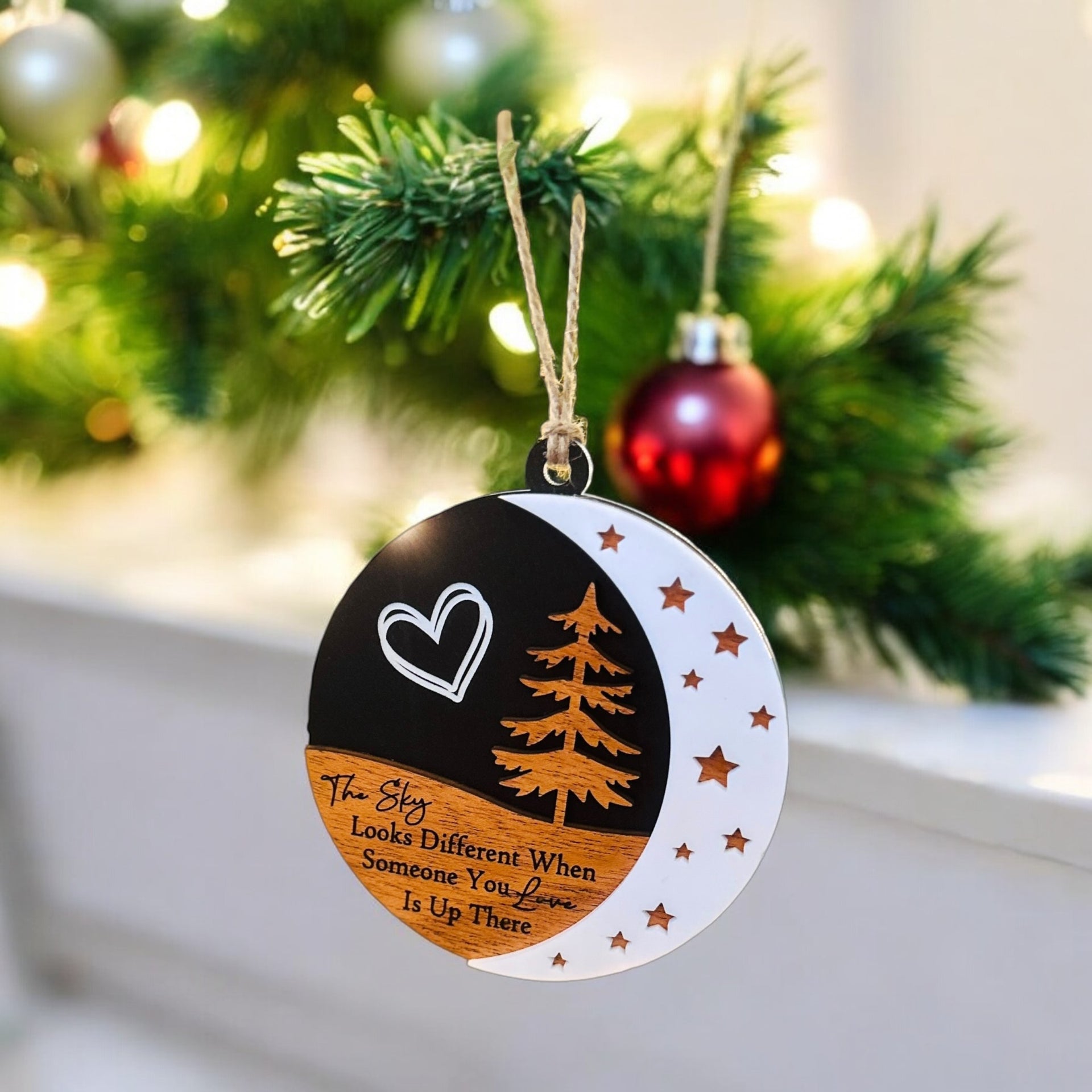 A round ornament hanging on a Christmas tree, featuring a heart symbol, a crescent moon, and stars, with the text "The sky looks different when someone you love is up there" printed on it.